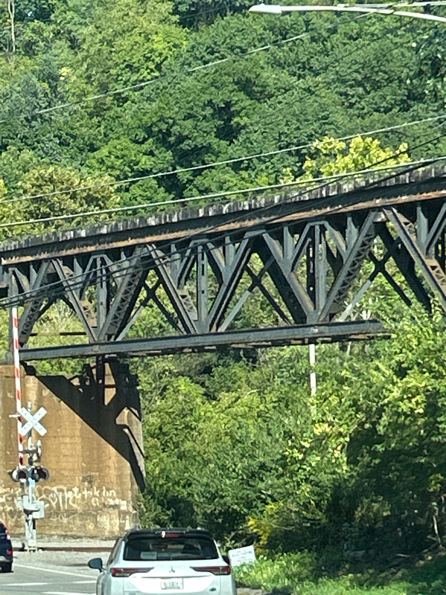 The image captures a view of a train bridge over a road, with lush green trees in the background. The bridge is a dark metal truss structure, with visible beams and supports. There are train tracks on top of the bridge. To the left of the bridge, a concrete structure is visible, with a railway crossing signal featuring red and white stripes and a white crossbuck sign. Below the bridge, a road is visible with two cars, one white SUV and a portion of a darker car on the left. The white SUV is positioned in the foreground, facing away from the viewer. The environment appears to be outdoors, with daylight casting shadows. The scene is located in Collier Township, United States of America.