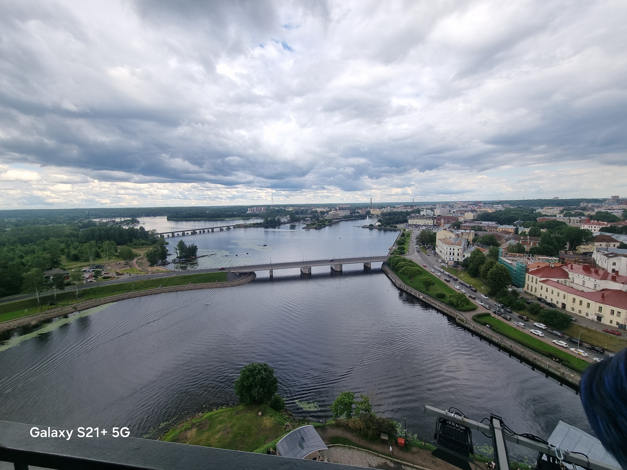 This is a high-angle, wide-shot aerial photograph of a city and a large body of water under a cloudy sky. The city is Vyborg, Russia. The scene captures the Vyborg Bay and the city's embankment.
In the foreground, a dark railing is visible along the bottom edge of the image, with the text "Galaxy S21+ 5G" in the lower left corner.
The middle ground is dominated by a wide, dark river with visible ripples on its surface. A long, concrete bridge with multiple arches spans the river from left to right, carrying a road with a few cars. On the left bank of the river, there is a green grassy area with trees and a road. On the right bank, a curved road runs alongside the river, lined with trees and a strip of green lawn. Beyond the road, a cityscape of buildings with red and light-colored facades is visible, interspersed with trees.
The background shows more of the city extending into the distance, with various buildings and more trees. A large, expansive sky filled with heavy, grey clouds takes up the upper portion of the image. The overall atmosphere is overcast, suggesting possibly gloomy or impending weather.