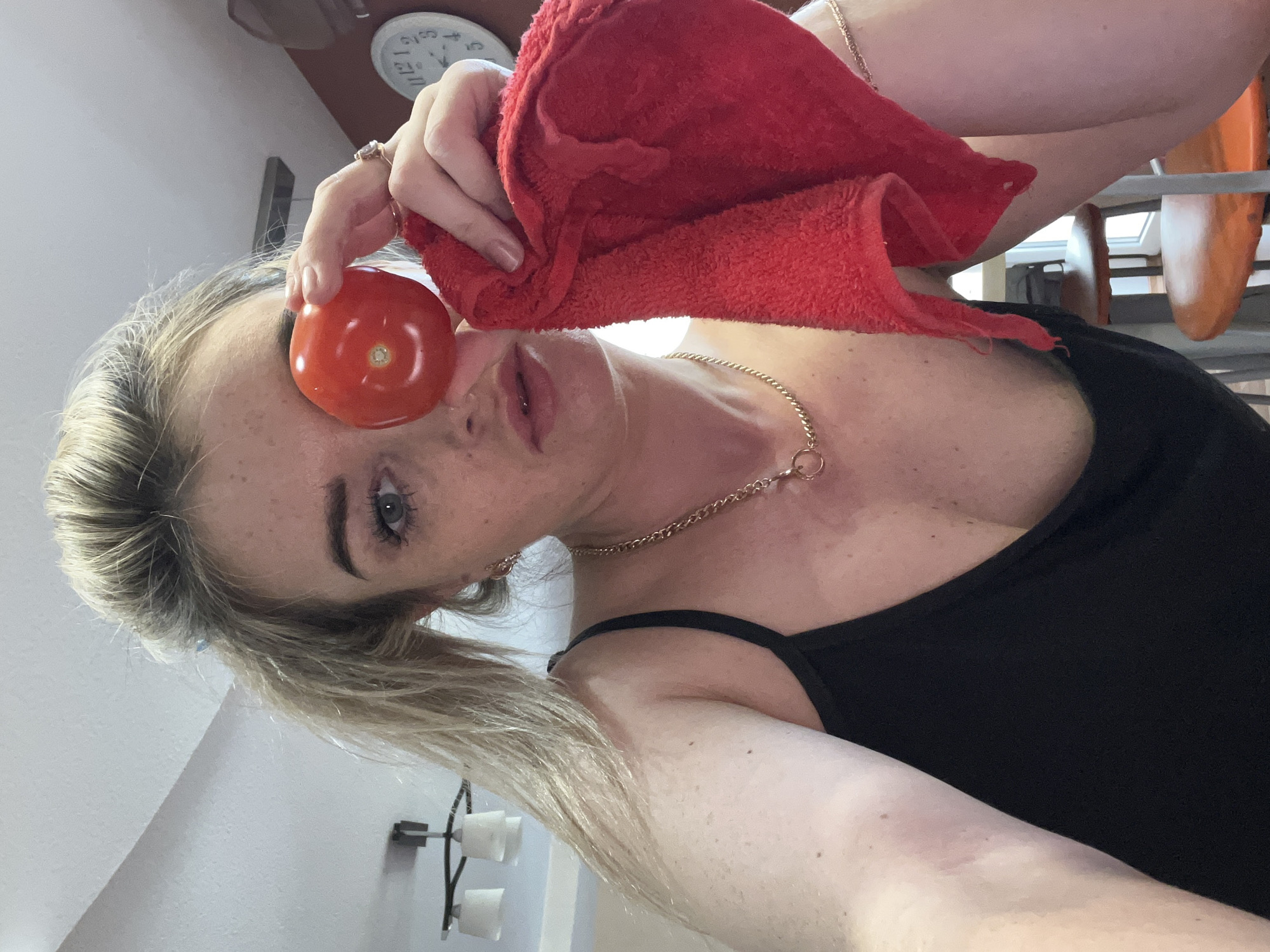A woman holds a tomato in front of her left eye and a red towel to the right of the tomato. She is wearing a black tank top and a gold necklace and ring. Her hair is blonde and pulled back in a ponytail. A clock is visible in the background. The location cannot be determined as Chaikovsky, Russia without additional information.