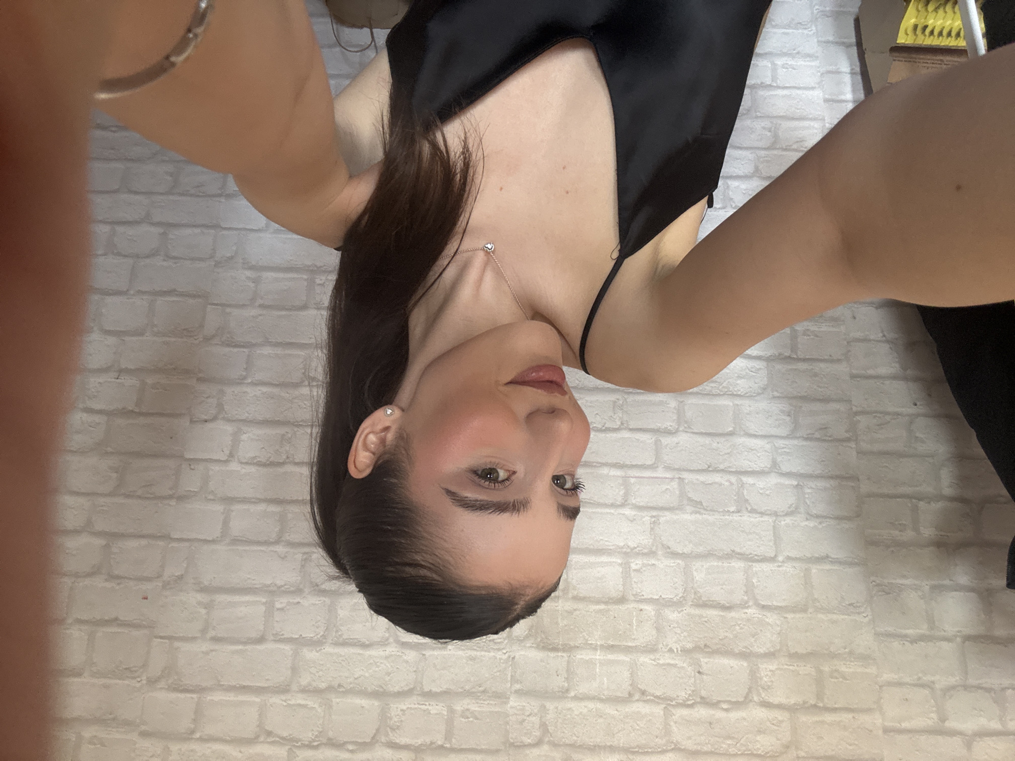 A photograph shows a woman with dark hair wearing a black dress, taken from a close, tilted, overhead angle. She wears a necklace and earrings. The background is a white brick wall. The location is Unknown, Unknown.