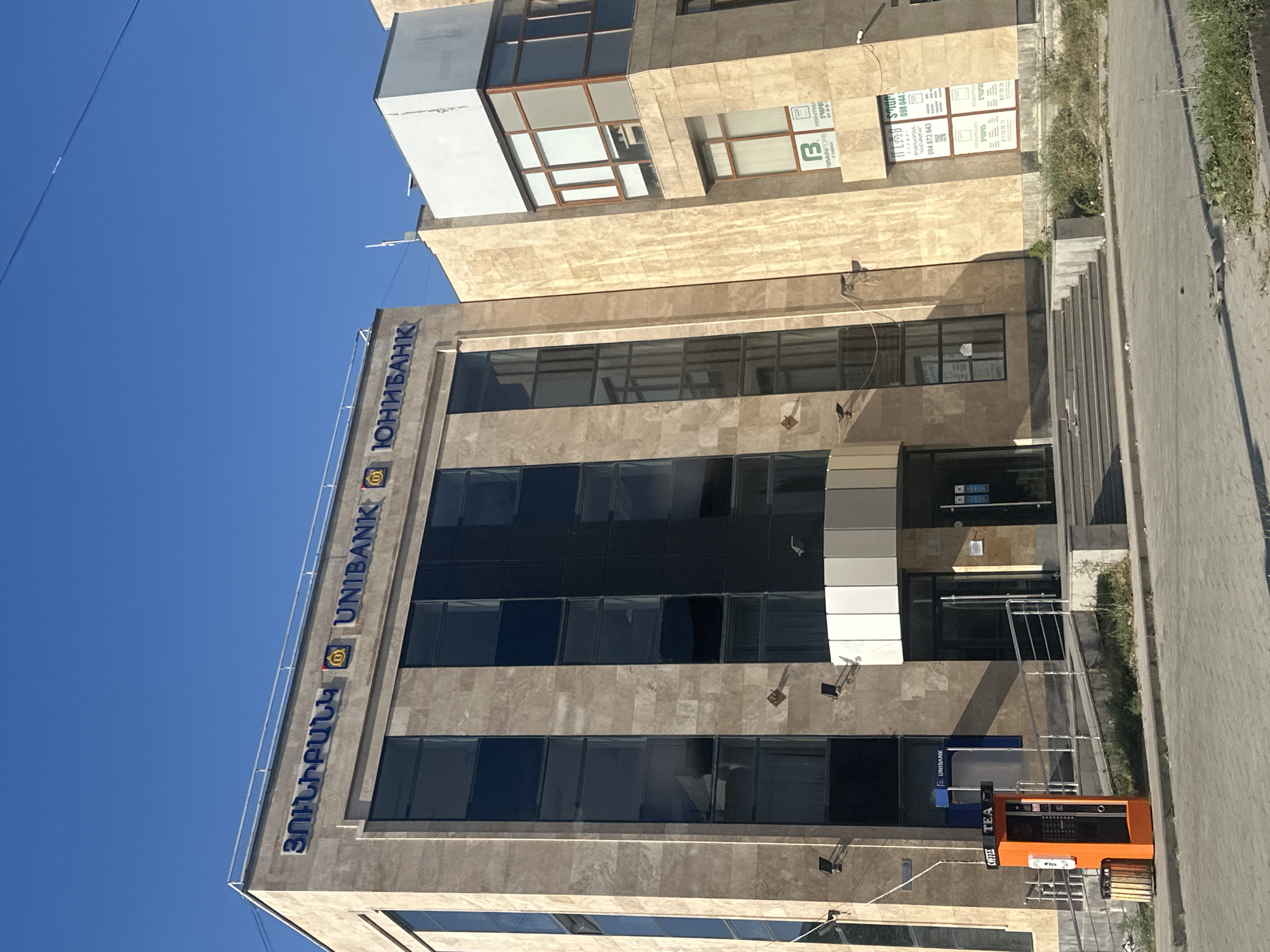 The image shows the exterior of a modern bank building on a clear day. The building, identified as "UNIBANK" in both Armenian and Russian script, is clad in beige marble and features large, dark-tinted windows. To the left of the main bank building is a smaller, connected structure with a white upper story and brown-trimmed windows.
In front of the bank, on the sidewalk, is an orange vending machine labeled "COFFEE TEA". A trash can is located next to the vending machine. A ramp and steps lead up to the entrance of the bank, with metal handrails. The environment appears to be a street in the city of Gyumri, Armenia, evidenced by the Armenian script on the building. The sky is a clear, bright blue, and the sun casts shadows on the building and the pavement. No people are visible in the image.