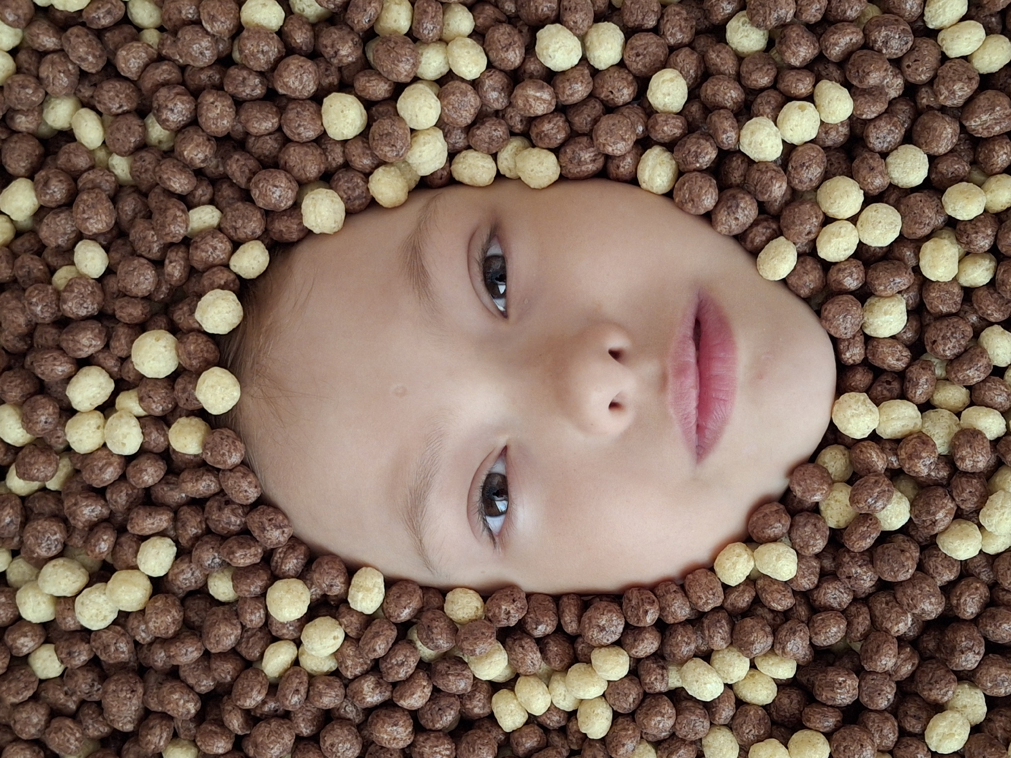 A close-up, eye-level shot depicts a child's face surrounded by cereal puffs of two colors: a light beige and a darker brown. The child is lying down, facing upwards. Their face is clean, with smooth skin, a small nose, and lips slightly parted. The child's eyes are open and focused on the viewer. The cereal puffs are densely packed around the face, creating a uniform texture in the background. The image was taken indoors. The location is not indicated by any objects or landmarks, so the city of Chaikovsky, Russia cannot be inferred.
