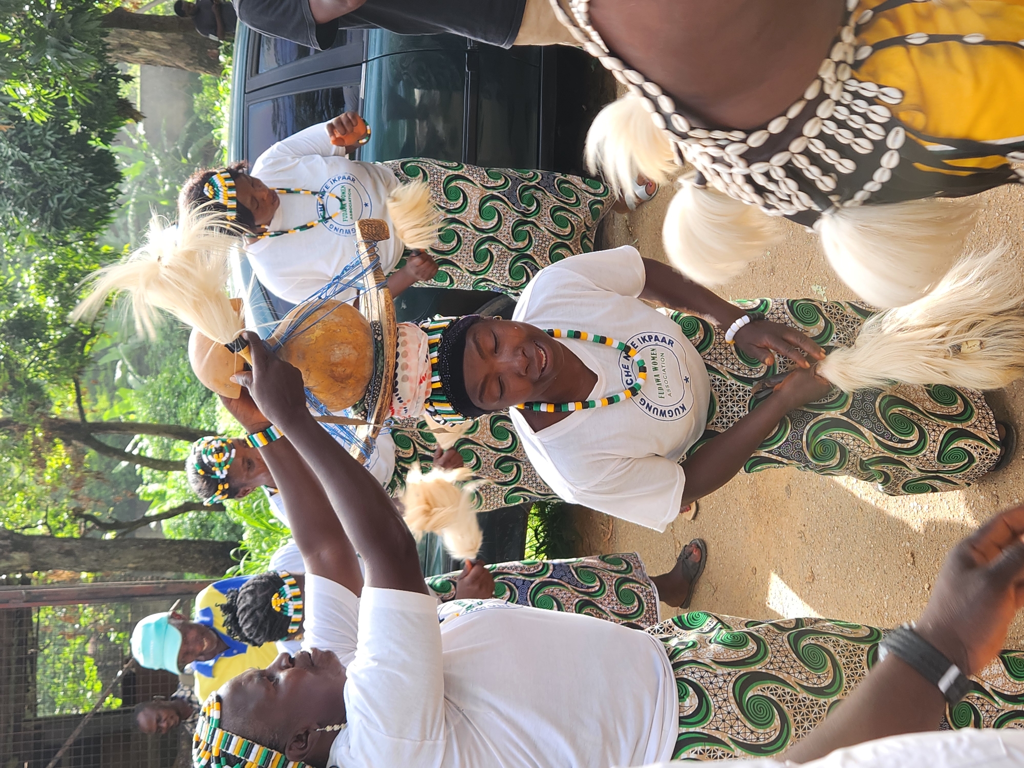 The image depicts an outdoor gathering in Jos, Nigeria, likely a cultural event or celebration. In the foreground, several women are dressed in traditional attire. One woman, in the center, wears a white t-shirt with a circular logo and a colorful beaded necklace. She is looking down and holding a fluffy white object. Another woman to her left, with a similar beaded headdress and necklace, is holding up a gourd on a wooden stand. Her white t-shirt also has a logo. Both women are wearing long skirts with a green and white swirling pattern. Behind them, a man in a light blue cap and a yellow and blue shirt is visible, smiling. In the background, a dark-colored vehicle is parked, and trees and foliage suggest a natural setting. The ground appears to be dirt or sand. Other individuals and details of the event are partially obscured.