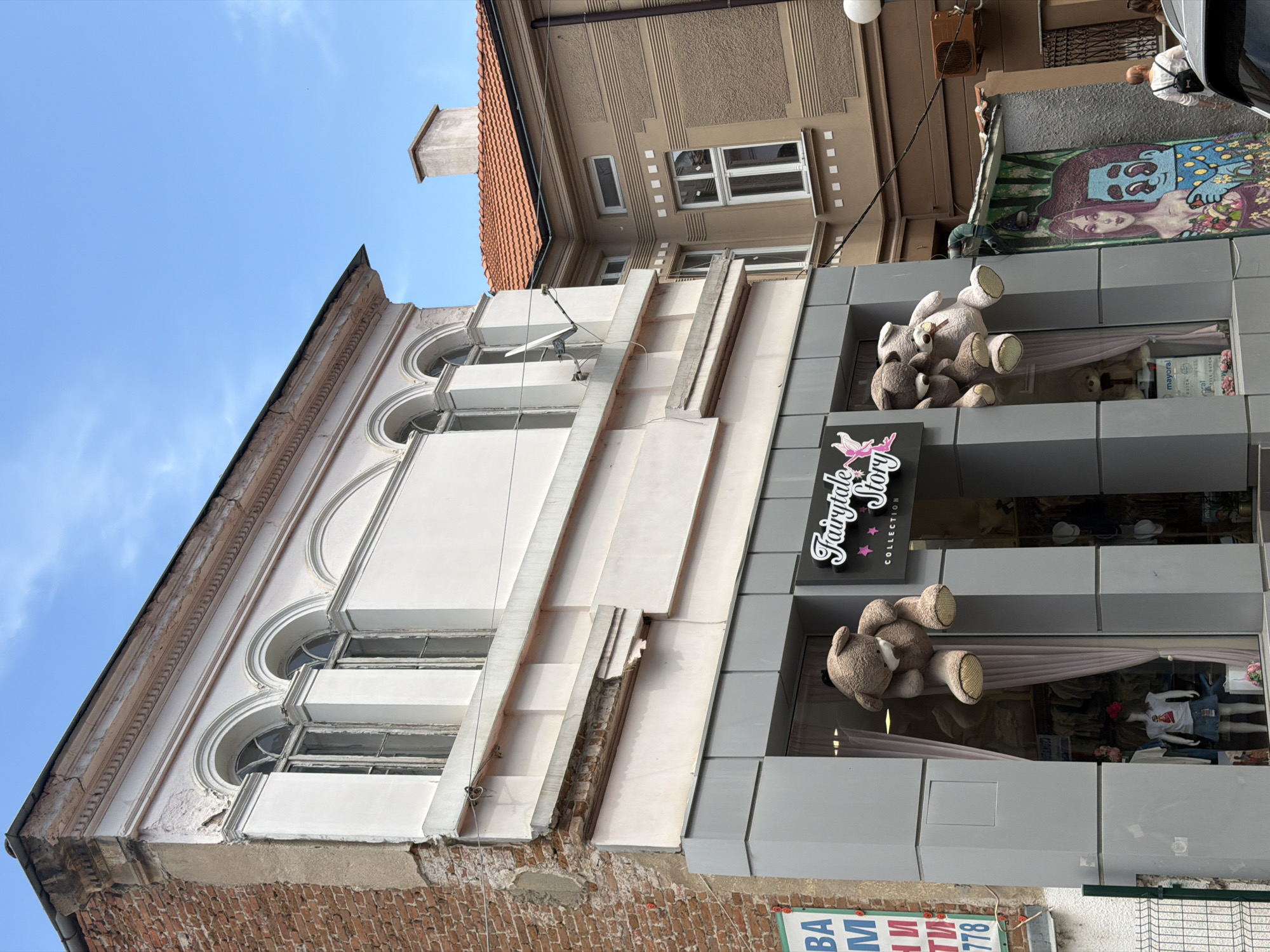 The image shows a building facade in Plovdiv, Bulgaria. The lower section features a brick exterior with arched windows. Above, there is a modern gray structure with two window displays. Each display contains large teddy bears, one above the other. A sign reading "Fairytale Story COLLECTION" is positioned between the window displays. The building adjacent to the gray structure has a brown exterior with white-framed windows and a red-tiled roof. In the upper-right corner, a mural is visible, and a person is standing nearby. A blue sky is visible in the upper left corner. A street sign with the numbers "778" is at the bottom of the image.