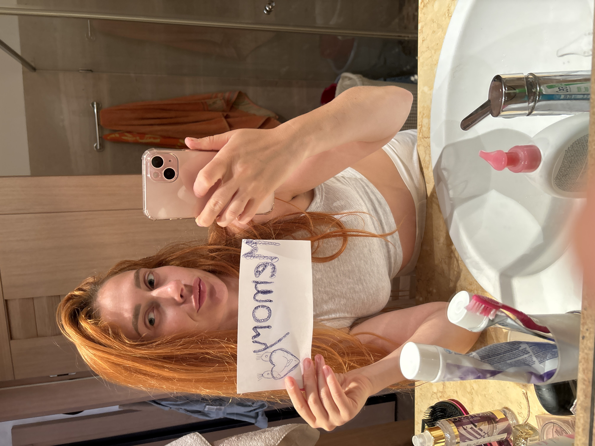 The image depicts an individual taking a mirror selfie within a bathroom in Budva, Montenegro. A young adult female with long, reddish-orange hair and light-colored eyes is visible, looking to her right with a slight head tilt and pursed lips. She is wearing a light-colored crop top. Her left hand holds a pink-cased smartphone, used to capture the image. Her right hand holds a white piece of paper displaying a mirrored inscription in dark blue marker that reads "Mommy" (or "Momom") accompanied by a heart drawing with small antennae-like protrusions.
The bathroom features a light-colored, possibly marble, countertop with a white ceramic sink and a chrome faucet. On the counter are various toiletries, including a white pump bottle with a pink dispenser, a container holding two toothpaste tubes (one with purple text, one with a red toothbrush inserted), a black hairbrush with a gold handle, and a clear spray bottle.
The background, seen in the mirror's reflection, includes light wood-paneled doors or cabinets on the left. A shower enclosure with clear glass panels and a chrome handle is visible, containing a chrome towel bar from which an orange-brown towel and a patterned towel hang. The shower walls are tiled in a light beige or brown hue. A light-colored garment is draped on a hook to the left, and a dark bucket or container is partially visible in the lower right background of the reflection.