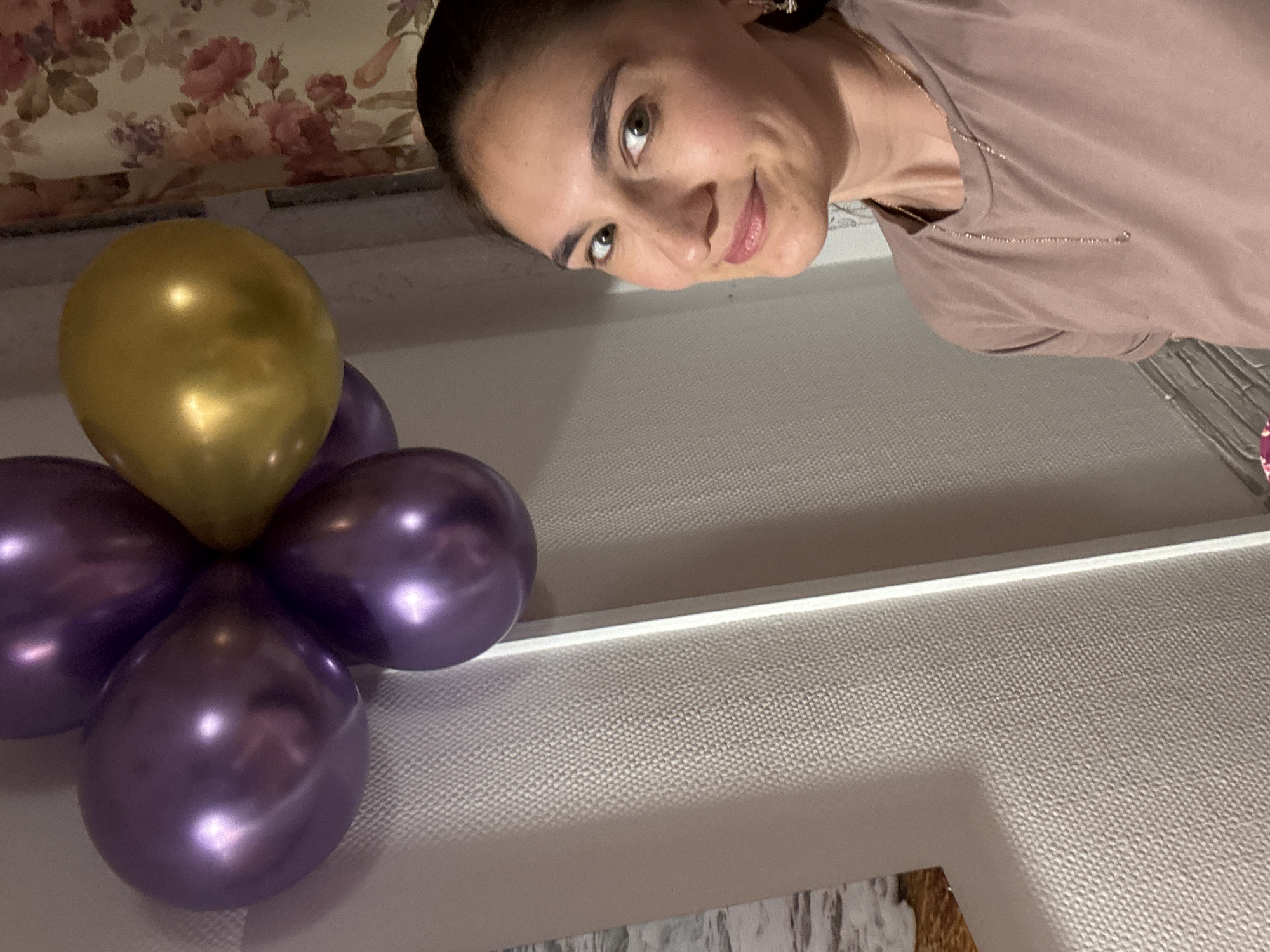 The image shows a woman with dark hair, wearing a light brown t-shirt and a gold chain necklace. Her face is turned towards the camera with a slight smile. In the foreground, there's an arrangement of balloons: one gold-colored balloon at the top and four purple balloons arranged around it, resembling a flower. The background includes a floral-patterned curtain and a textured white surface, likely a wall or ceiling. Part of a picture frame with a blurred image is visible at the bottom. There's no specific indication of the location being Sochi, Russia, so this cannot be confirmed from the image.