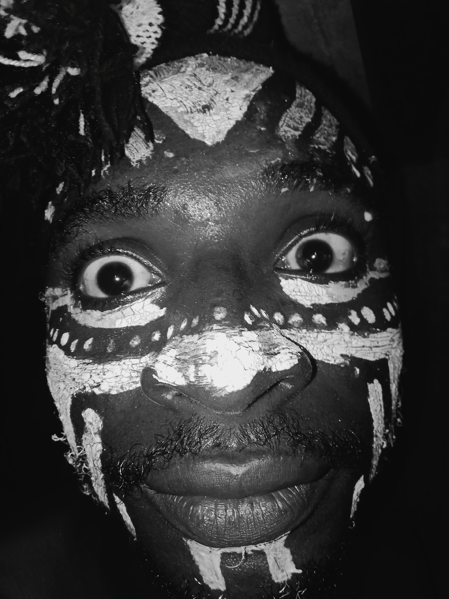 A close-up, black and white photograph captures a person's face adorned with tribal-style face paint. The paint is primarily white, contrasting sharply with the darker skin tone. The designs are elaborate, featuring patterns of lines, dots, and geometric shapes around the eyes, forehead, nose, and mouth. The individual's eyes are wide and focused directly at the camera, creating an intense gaze. The mouth is slightly open, suggesting a slight smile. The background is dark and indistinct, placing the emphasis entirely on the face. The texture of the paint appears cracked in places, giving the impression that it has been applied for some time. The scene is in the city of Unknown, Unknown.