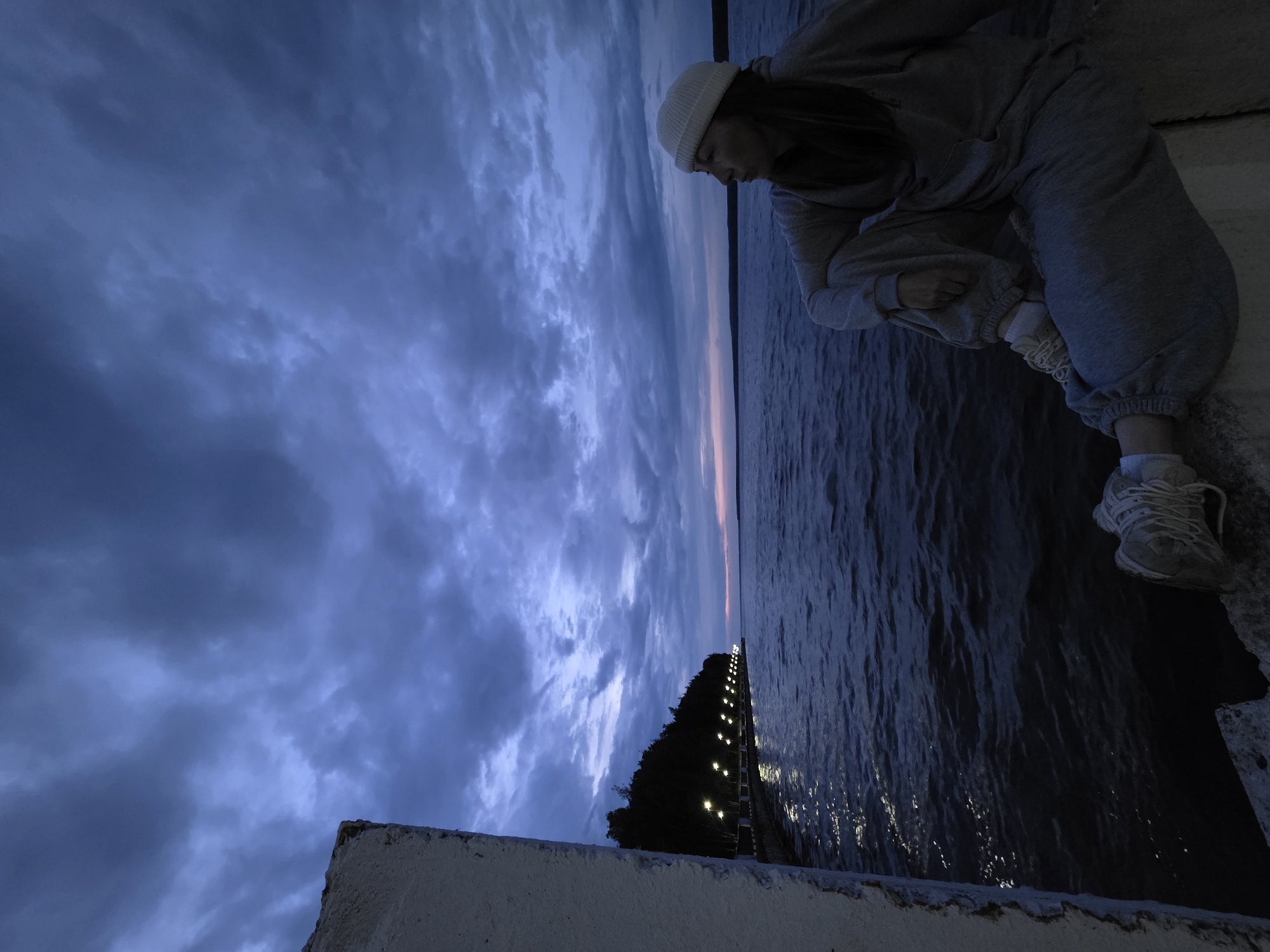 A girl in a gray sweatsuit and a white knit cap is crouching on a concrete ledge, looking down at the water. The body of water appears to be part of the Kama River near the city of Chaikovsky, Russia. The sky is dark and cloudy, suggesting that it's dusk or dawn. In the distance, there's a line of lights that could be part of a bridge or embankment. The lighting in the photograph indicates that it is in the evening. The concrete ledge is in the foreground, and the girl is positioned to the right, with the water and sky filling the rest of the frame.
