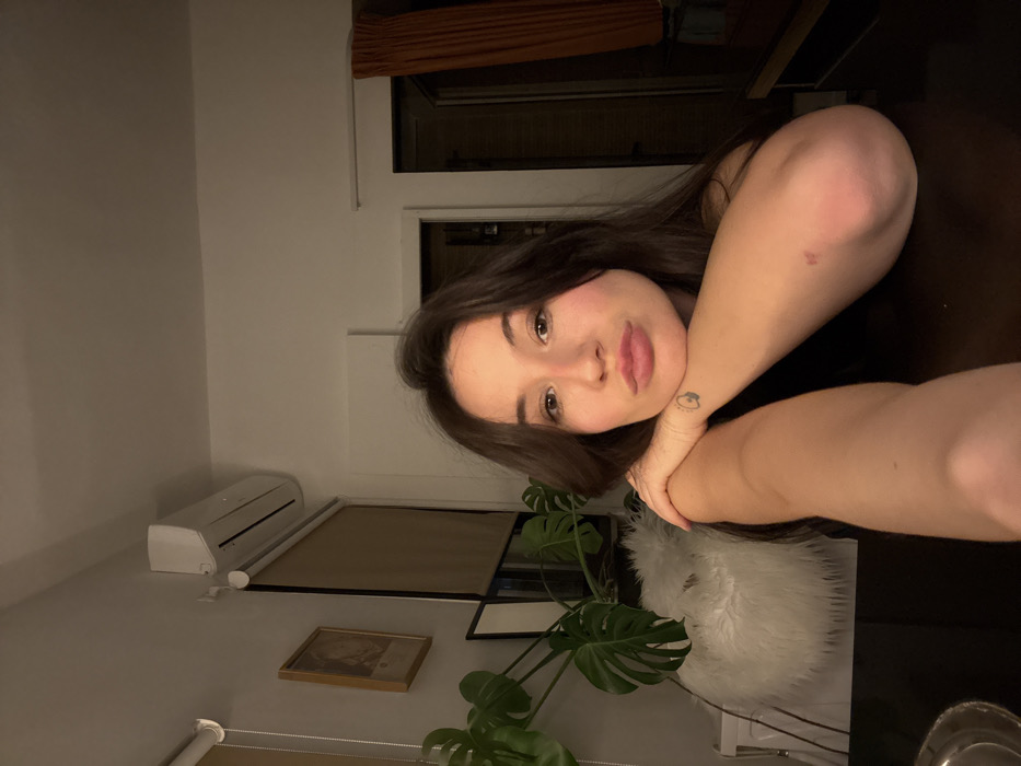 An indoor scene within a residential apartment, likely located in Tel-Aviv, Israel, is depicted under artificial, subdued lighting. A female individual, positioned centrally, gazes directly at the camera with pursed lips, her head resting on her forearms which are placed on a dark, polished table surface. She has long, dark hair and a fair complexion. A small, dark, abstract tattoo is visible on her left wrist, and a reddish mark is present on her right elbow. She is wearing a dark-colored garment.
The background features light-colored walls. In the upper left quadrant, a white split-unit air conditioner is mounted on the wall. Below it, a window is covered by light brown roller blinds. To the immediate left of the blinds, a framed artwork hangs on the wall. A large green leafy plant, resembling a Monstera, is visible behind the subject's left side, next to a white, furry object. To the right of the subject, a doorway or opening is partially obscured by dark orange-brown curtains, revealing a dimly lit interior beyond.