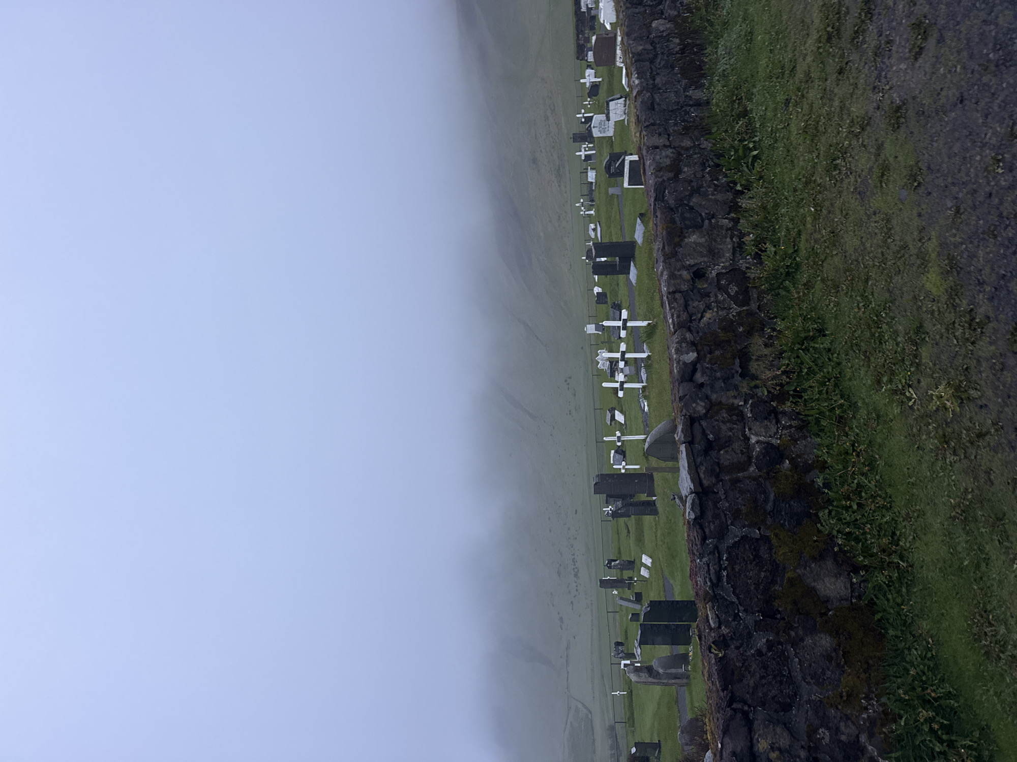 This image depicts a cemetery in Vik, Iceland, on a foggy day. The landscape is characterized by a grassy field that slopes upward into the fog. Scattered across the field are numerous headstones and crosses, indicating grave markers. A dark stone wall runs along the right edge of the frame, separating the cemetery from a path or road covered in grass and gravel. The fog obscures the distant features, creating a somber, ethereal atmosphere. There are no people visible in the frame.