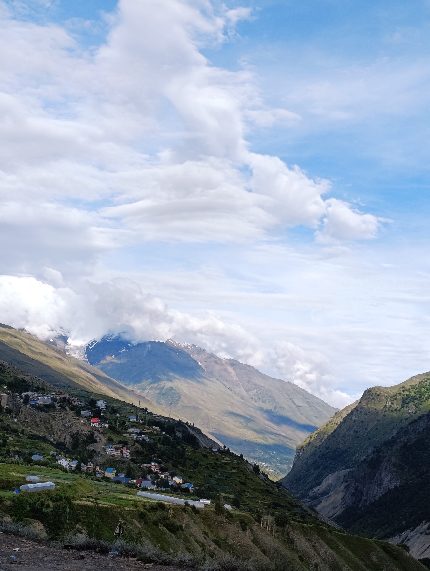 The image depicts a mountainous landscape in the vicinity of Gondhla, India, under a sky with scattered white clouds. In the foreground, a steep, vegetated slope is visible, with terraced fields and several small white structures, possibly greenhouses, scattered across it. A small village with numerous houses, some featuring red roofs, is built on the hillside further up. Beyond the village, majestic mountains rise, with some peaks covered in snow and shrouded in clouds. The sunlight casts dramatic shadows across the slopes, highlighting the rugged terrain. The overall scene is one of a remote, rural mountainous settlement.