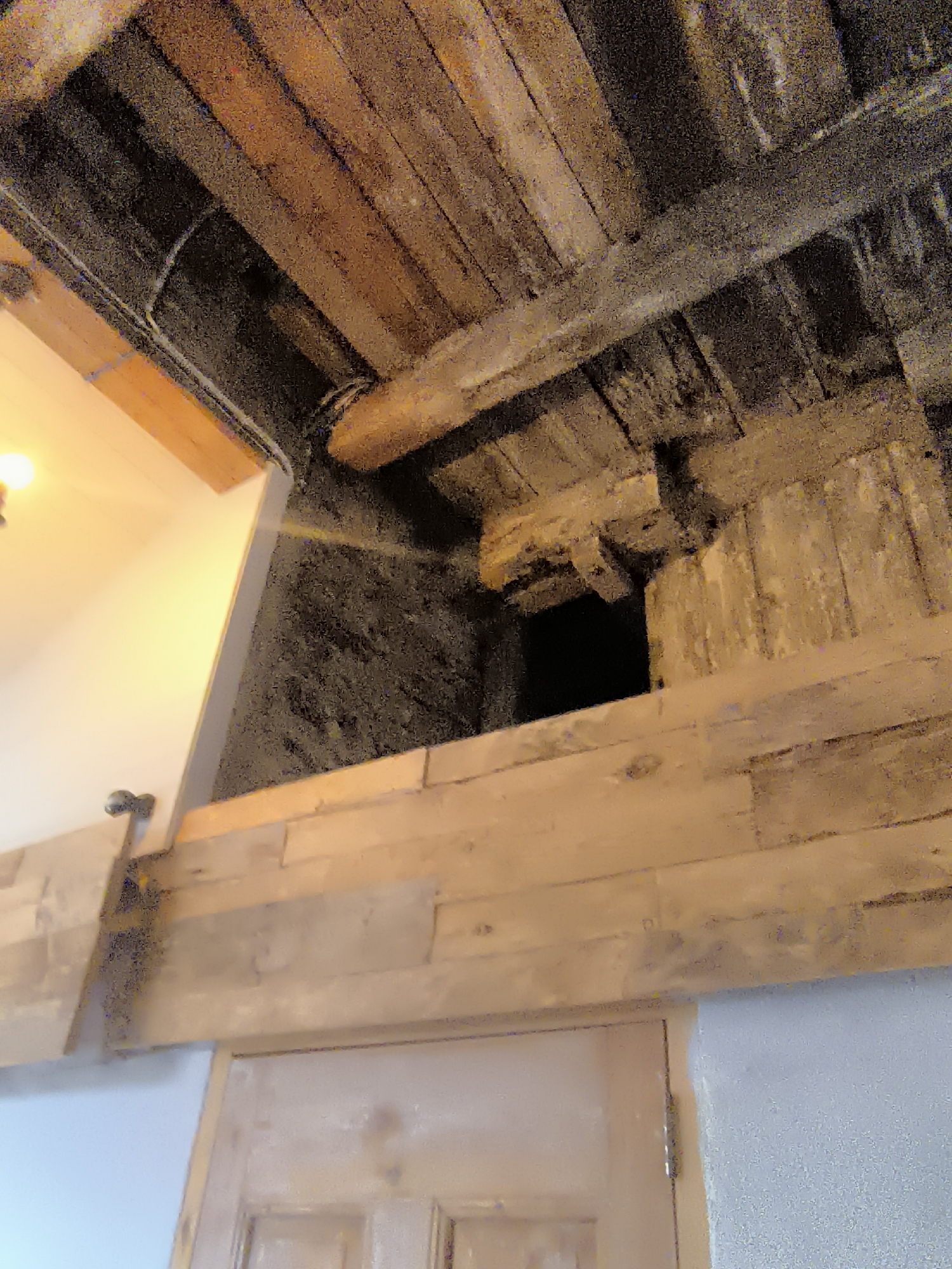 The image shows a dark, narrow opening between wooden beams and stone walls, suggestive of an attic or crawl space. A light fixture is visible to the left, illuminating part of a white wall. The overall impression is one of aged construction and a utilitarian space. The location in Kuldīga, Latvia, is not visually discernible from the image itself; it is an external piece of information.
