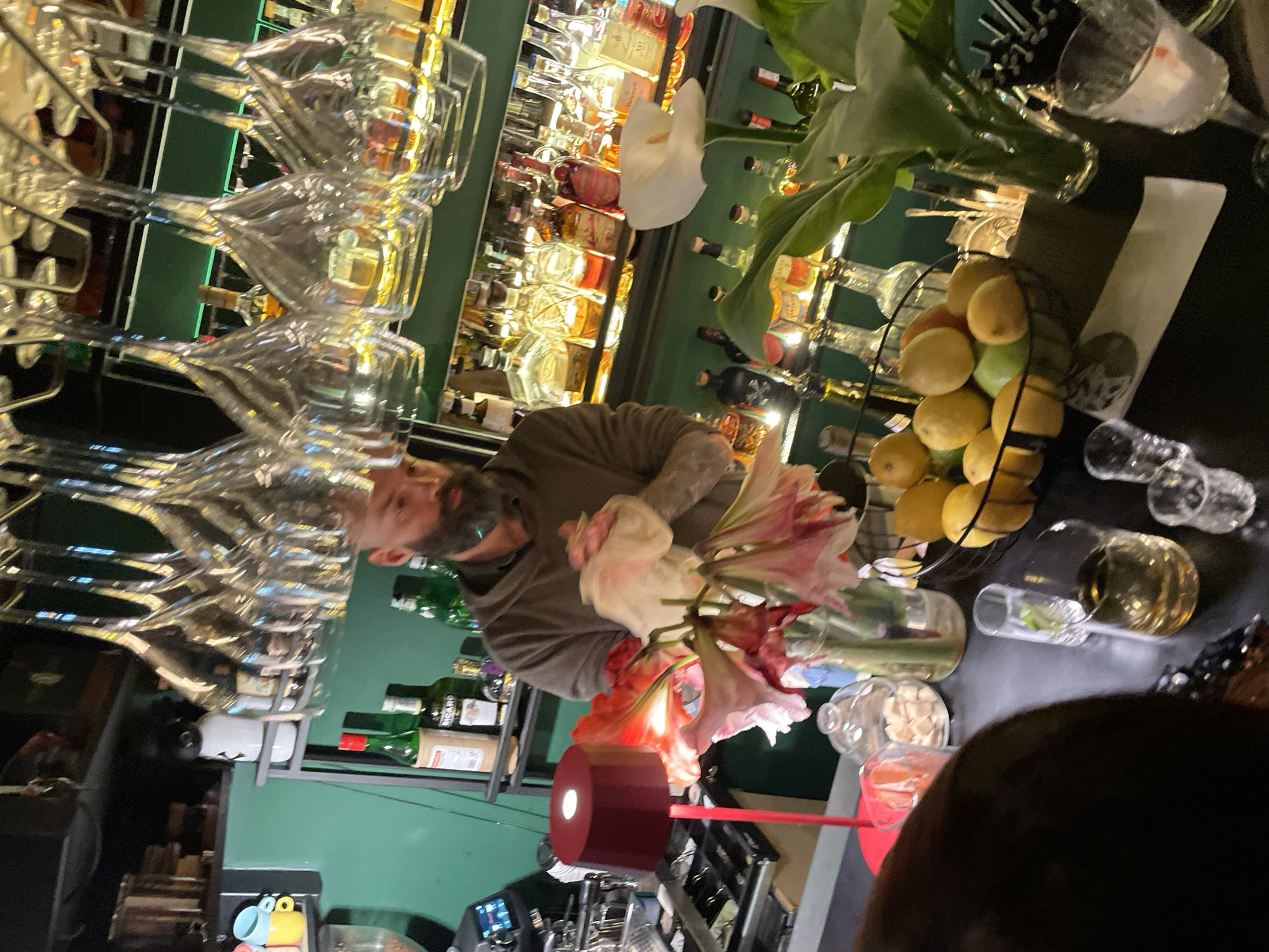 The scene captures the interior of a bar, likely in Budva, Montenegro. A man with a beard and tattoos is partially visible, positioned between a liquor shelf stocked with bottles and an array of wine glasses hanging overhead. In the foreground, a table features a decorative arrangement of lilies in a vase, a bowl of citrus fruits, a golden-colored liquid in a glass, and several small glasses. A person's head is partially in the frame, suggesting a close-up view of the bar's setting.