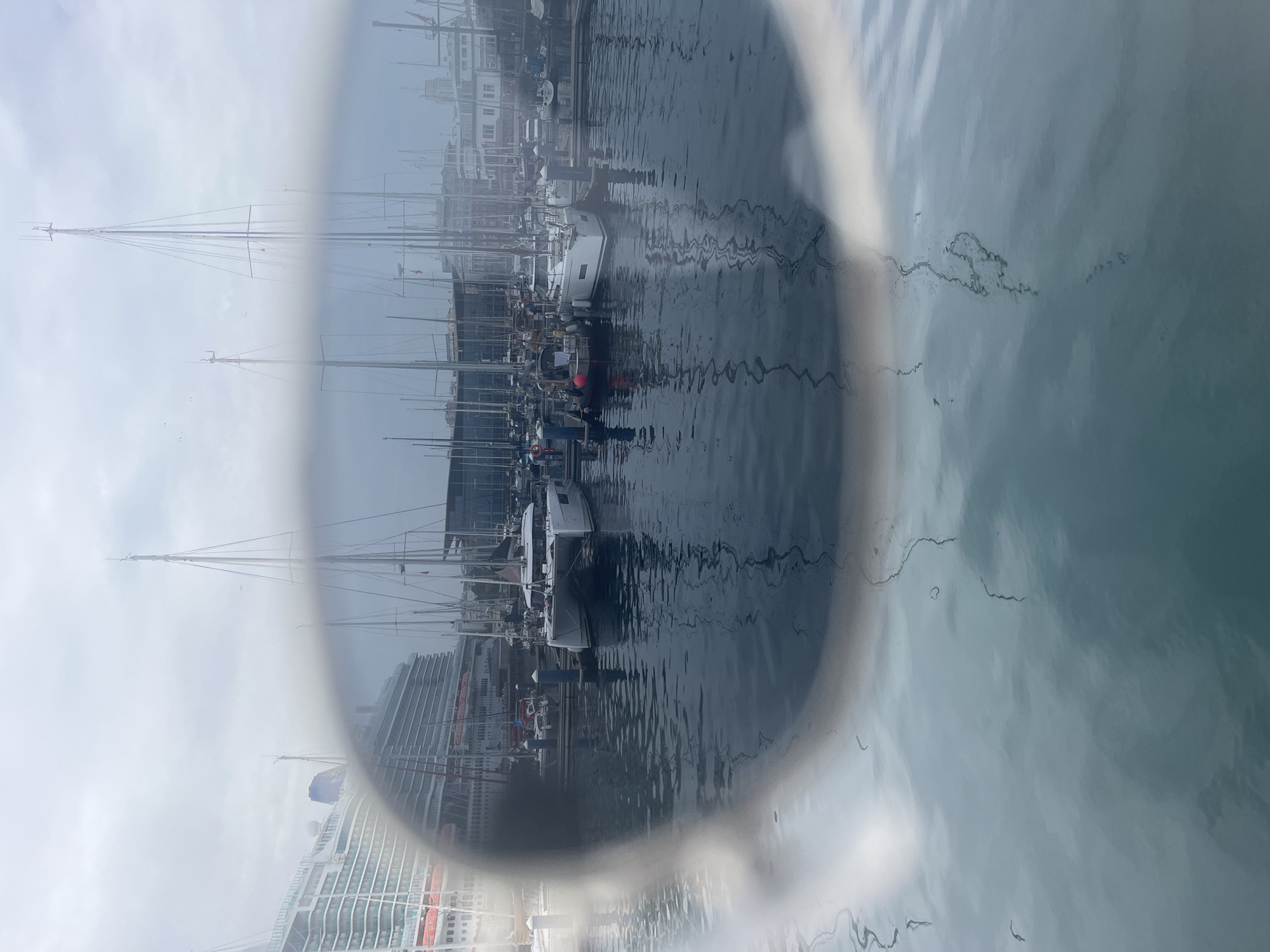 This image captures a harbor scene in A Coruña, Spain, with a large cruise ship docked on the left and numerous sailboats moored in the foreground. The view is partially obscured by the reflection or lens of sunglasses, creating a blurred, vignetted effect. The water is calm, reflecting the overcast sky and the masts of the sailboats. Buildings line the waterfront in the background, some appearing to be modern structures. The overall atmosphere is subdued, consistent with a cloudy day. No people are visible in the image, and the focus is on the maritime elements of the harbor.