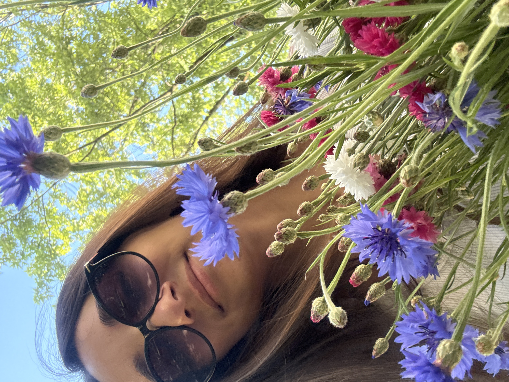 A low-angle, outdoor, close-up shot shows a young woman holding a bouquet of cornflowers and other wildflowers. She is wearing large, black sunglasses, and her long, brown hair is visible. The woman is smiling slightly, and her face is partially obscured by the flowers. The background consists of lush green foliage and a clear blue sky, suggesting a sunny day in a park or garden. The flowers are vibrant, with shades of blue, pink, and white. The scene evokes a sense of joy and natural beauty. The specific location in Kyiv, Ukraine, is not discernible from the image itself.