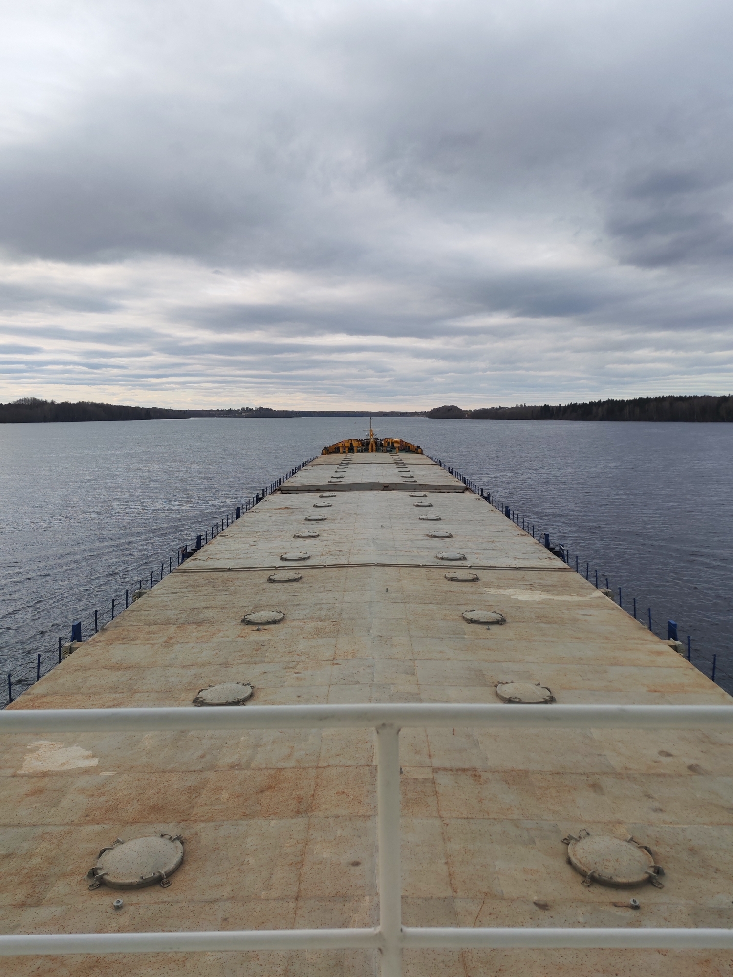 The image shows a view from a ship's deck on a body of water, likely a river or a wide lake, under a cloudy sky. The deck is tan and marked with several round, bolted hatches. A white railing is visible in the foreground. Across the water, forested land is visible. The scene is taken in Khoroshevo, Russia.