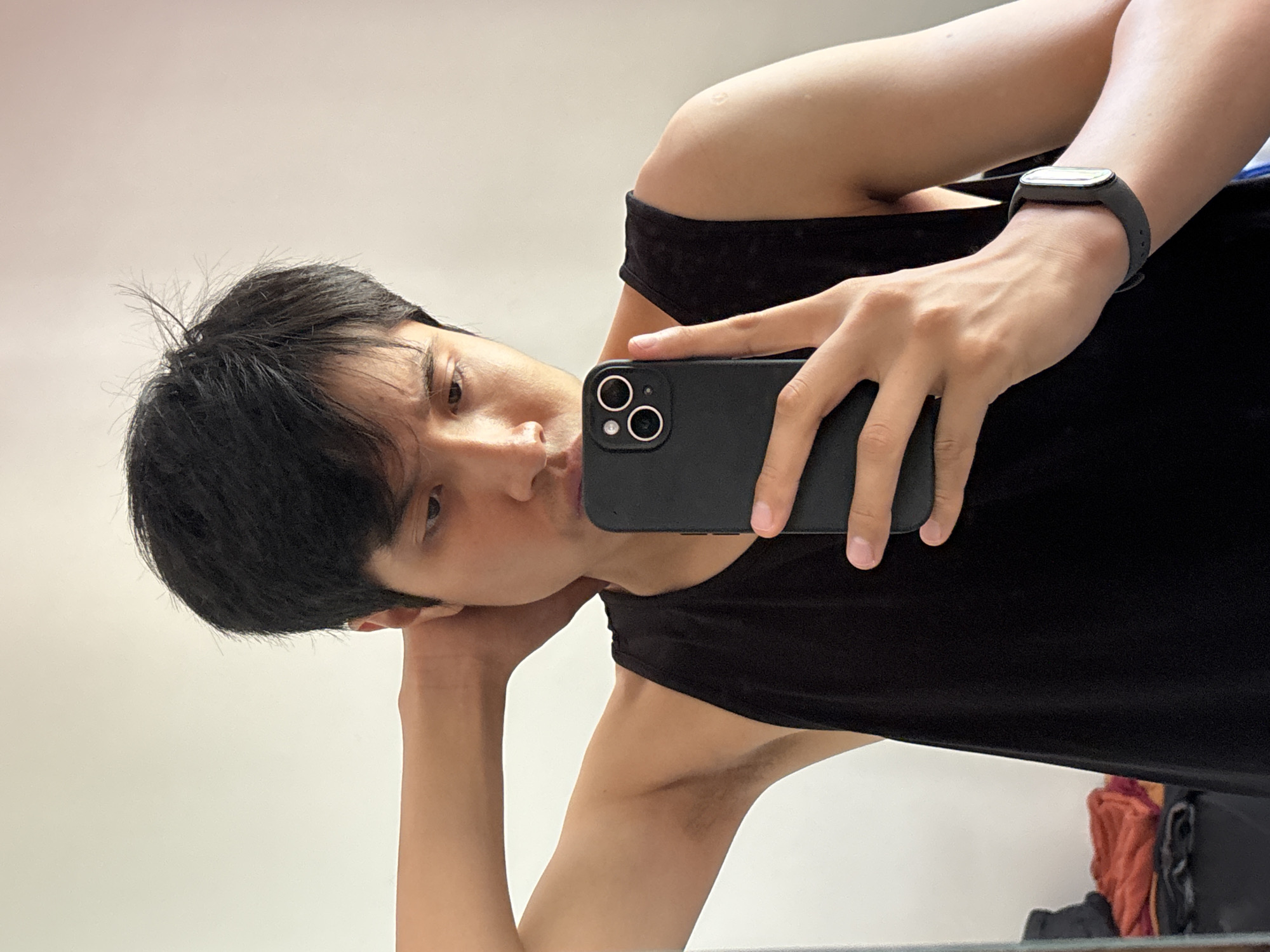 A person with dark hair and light skin is taking a selfie with a black smartphone. The person is wearing a black tank top and a black smartwatch on their left wrist. They are leaning on their left arm, which is visible. The background is a plain white wall. The location cannot be determined from the image.