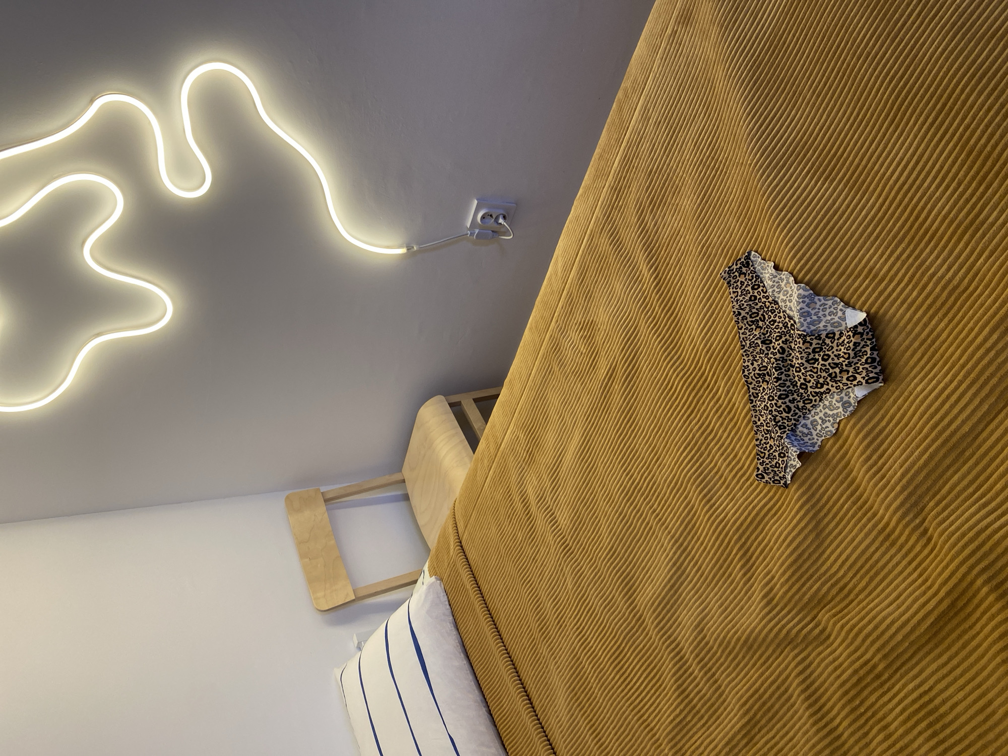 In a room with light gray walls, a bed covered with a mustard-yellow corduroy bedspread dominates the foreground. A white pillow with blue stripes lies at the head of the bed. A wooden chair with a simple design is positioned behind the bed. On the bedspread, a pair of leopard-print underwear is casually placed. A white neon light fixture with an abstract shape is mounted on the wall above the bed. An electrical outlet with a cord connected to the light is visible on the wall. The scene appears to be in a bedroom, possibly in a home or hotel. There is no information to suggest this location is in Izhevsk, Russia.