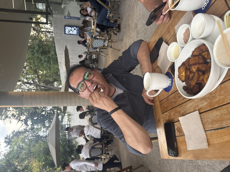 The image depicts an outdoor dining scene at Proscenio Cafe, located in Mexico City, Mexico. In the immediate foreground, a man with dark hair and black-rimmed glasses, wearing a dark blue long-sleeved shirt, is seated at a wooden slatted table. He is actively consuming food, holding a piece of fried plantain with a white topping to his open mouth, with a playful expression. On the table to his right is a white mug resting on a blue coaster. A black smartphone is placed face-down to his left, adjacent to a brown paper napkin. Central on the table is a white oval bowl filled with numerous fried plantain slices. This is accompanied by smaller white ramekins containing white cream, reddish-brown sprinkles, and a light-colored liquid (possibly condensed milk or syrup). Partially visible on the far right are another individual's hands holding a black smartphone, with a white mug and a straw also present. The background shows an active outdoor cafe area with multiple patrons seated at tables featuring bistro-style chairs, many positioned under large beige umbrellas. Several staff members, dressed in white shirts and grey aprons, are visible attending to guests. Lush green trees occupy the distant background, and a modern concrete building, displaying the illuminated "PROSCENIO CAFE" sign, forms part of the cafe structure. A prominent cylindrical concrete pillar stands in the mid-ground. The lighting suggests either late afternoon or an overcast daytime sky.