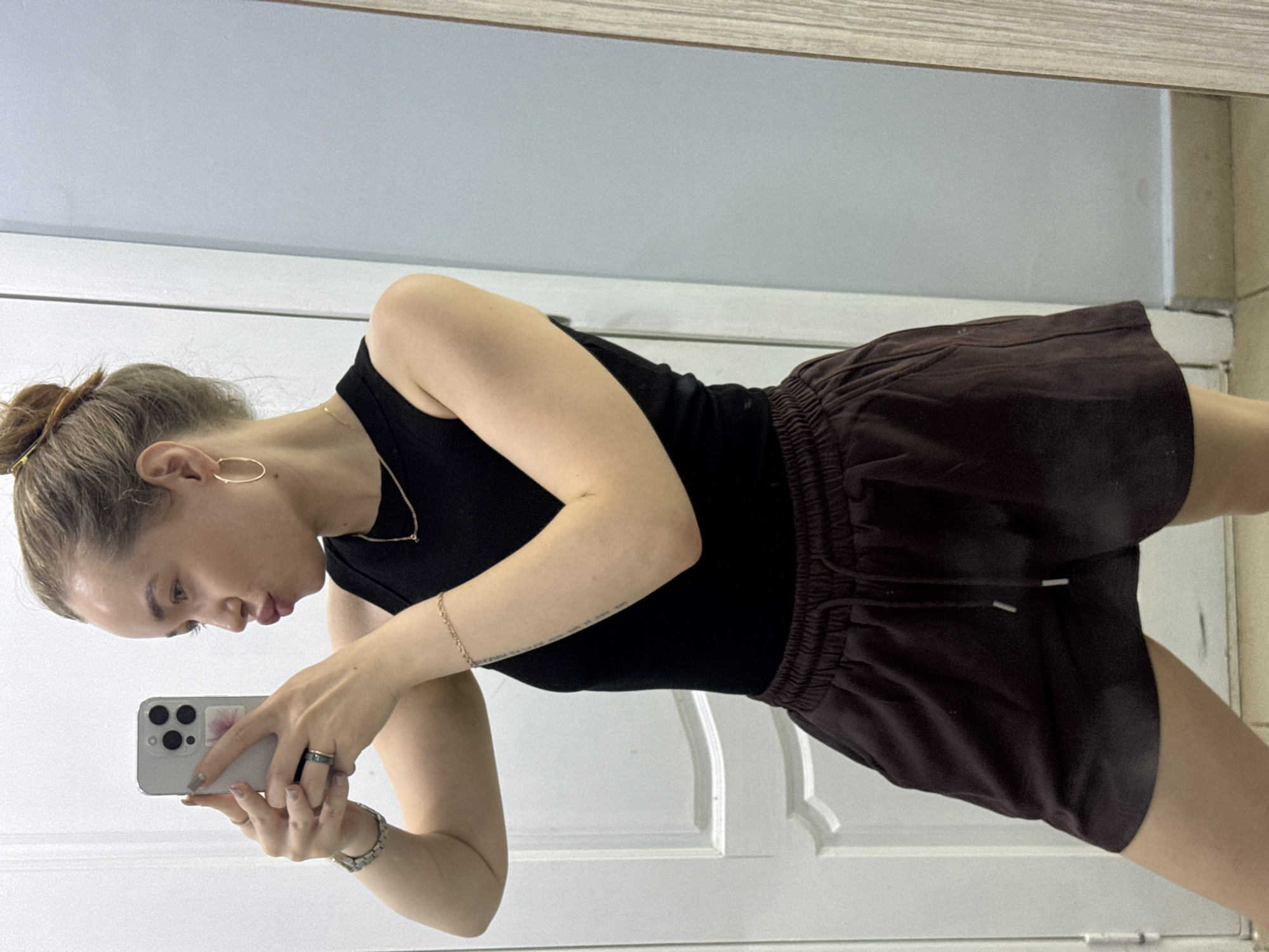 A woman is taking a selfie in front of a mirror. She has her hair up in a bun and is wearing hoop earrings, a gold necklace, a black tank top, and brown shorts. She is holding an iPhone in her left hand. The background is a white door and a grey wall. There is a small section of wall with green tiles in the top right corner of the picture. It is impossible to determine whether the photograph was taken in the city of Chaikovsky, Russia.