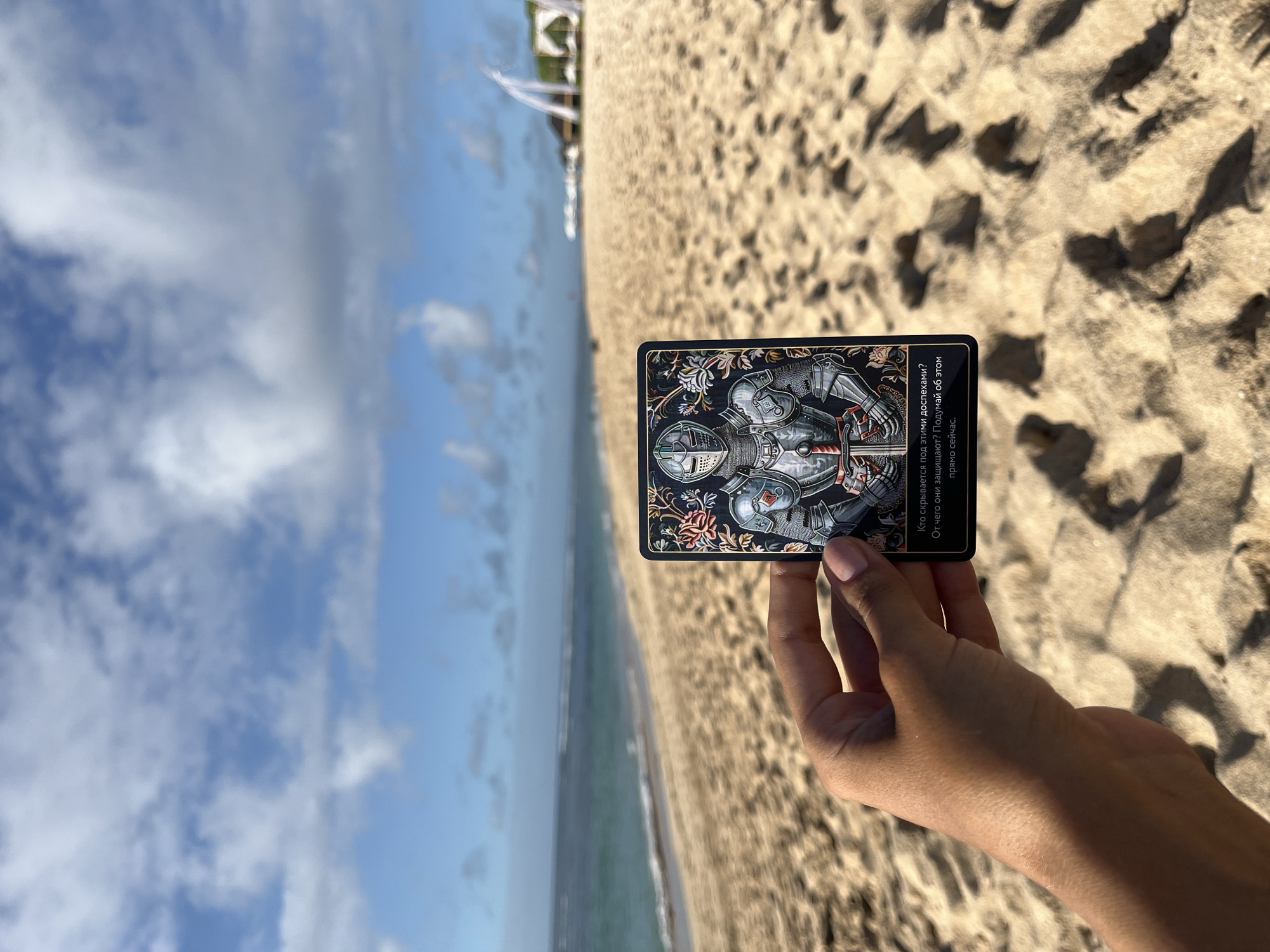 A person is holding a tarot card in their hand on a sandy beach. The backdrop is a beach with a blue sky dotted with clouds and the ocean meeting the shore. The beach stretches into the distance, with blurred structures in the far background. The tarot card features an illustration of a knight in armor with a sword, surrounded by ornate floral designs. The text on the card in Cyrillic reads "Кто скрывается под этими доспехами? От чего они защищают? Подумай об этом прямо сейчас." which translates to "Who is hiding under this armor? What are they protecting from? Think about it right now." The location is Kuta Selatan, Indonesia.