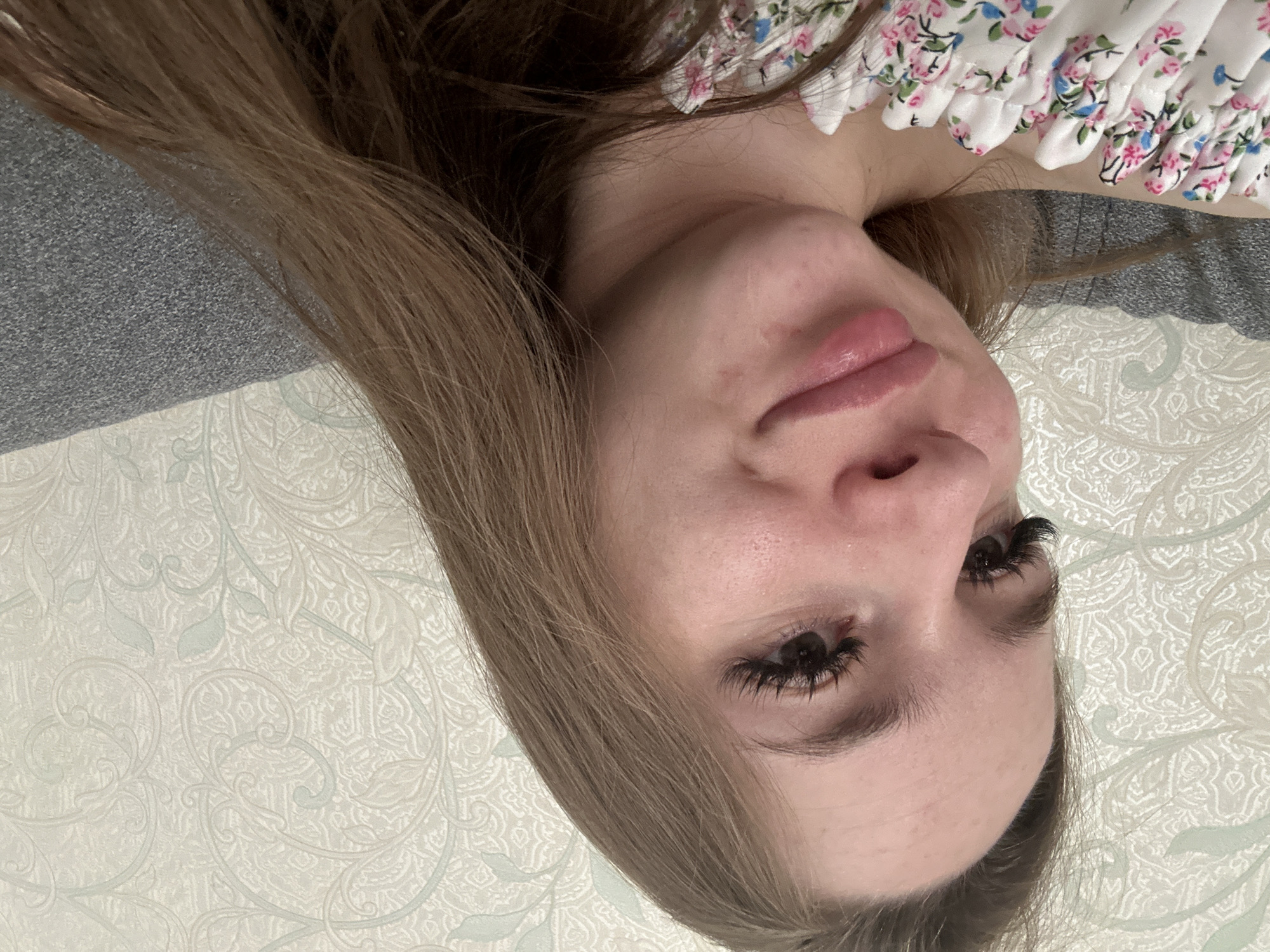 The primary image displays a close-up of a person, likely female, lying down and looking towards the camera from an inverted perspective. Her light brown hair cascades around her face. She is wearing a white top adorned with a small floral pattern in shades of pink, blue, and green.
Her face is visible, featuring dark, full eyelashes, a small nose, and plump, pink lips. There are a few minor blemishes on her skin, particularly around her chin and cheek. Her expression is neutral, possibly with a hint of a soft smile.
The background consists of two main elements: a grey, textured fabric, which could be clothing, a blanket, or a piece of furniture; and a lighter patterned surface, likely wallpaper or bedding, featuring intricate, light grey and white swirling floral and leaf designs. The combination of these elements suggests an indoor setting, possibly a bedroom or living room.
No specific activity is taking place beyond posing for the photo. The lighting is soft and does not indicate a specific time of day. There is no visible text in the image.