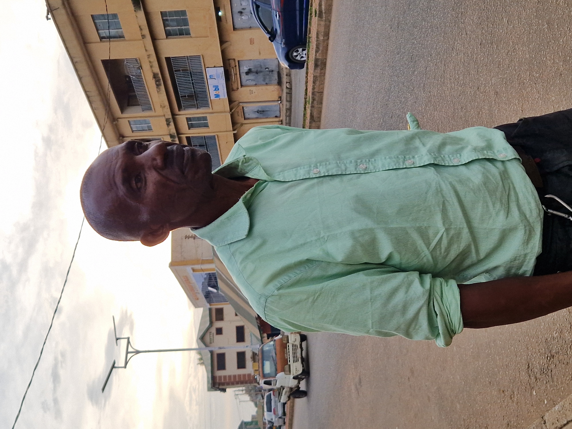 A dark-skinned bald man with a light green collared shirt stands on a paved road in Kabong, Nigeria. The shirt is buttoned up and the sleeves are rolled to his elbows. He is looking to his right with a neutral expression. Behind him, a yellow two-story building with balconies and windows is visible. A blue car is parked on the right side of the road. In the background, there is a white and brown truck on the left side of the road, along with other vehicles and buildings. A street lamp with two lights is also present. The sky is overcast with clouds. The scene appears to be a street in a town.