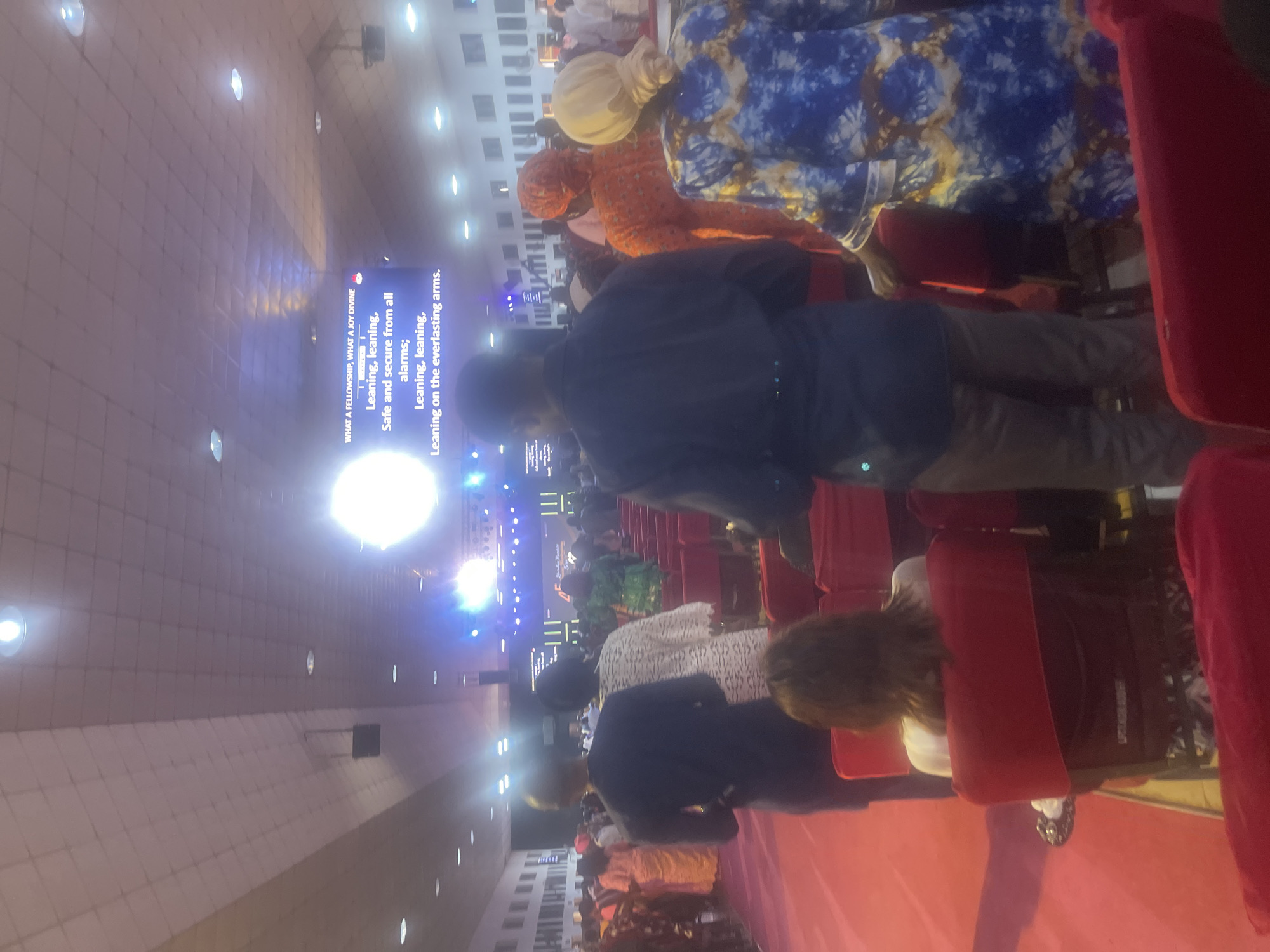 The image shows a gathering in a large hall, possibly a church or event center in Abuja, Nigeria. People are seated in red chairs facing a stage with a screen displaying text: "WHAT A FELLOWSHIP, WHAT A JOY DIVINE" followed by lyrics about leaning on everlasting arms. Some attendees are wearing traditional Nigerian clothing, including headwraps. The hall has a high ceiling with multiple lights.