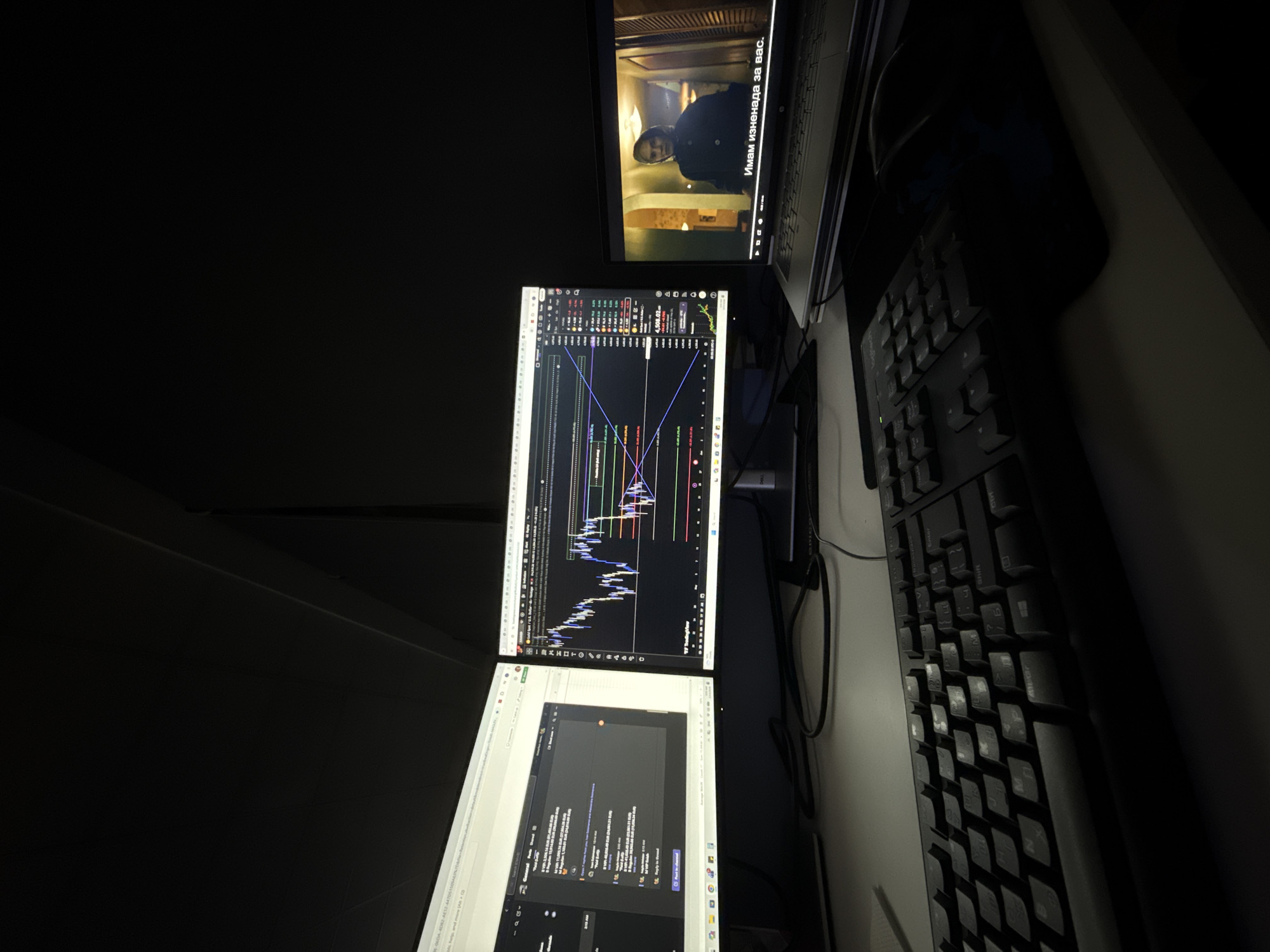 The image shows a dimly lit workspace. There are three monitors stacked vertically. The top monitor displays a video with Bulgarian subtitles. The middle monitor shows a stock trading chart with technical analysis lines. The bottom monitor displays a computer program. To the right of the monitors are two keyboards, arranged vertically. The environment is dark and the overall scene appears to be a workstation. No people are visible. There is no indication of the location in the city of Plovdiv, Bulgaria.