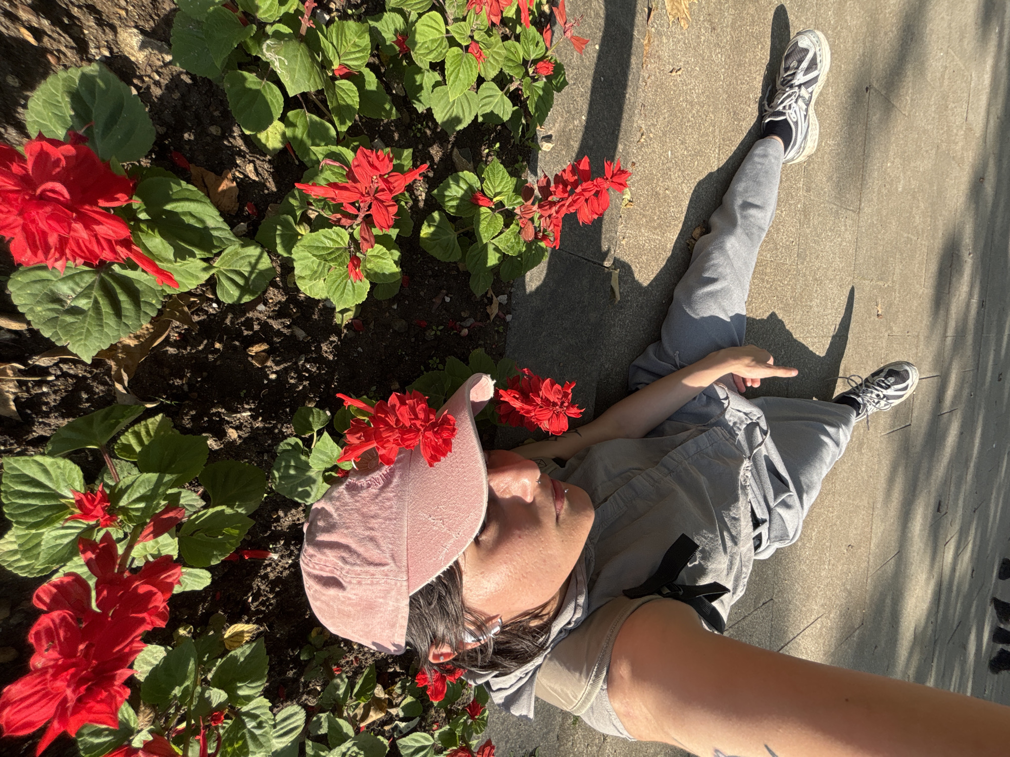 The scene depicts an outdoor urban or park environment under direct sunlight, featuring a paved area adjacent to a flower bed.
One individual is lying supine on a light-colored paved surface, positioned with their head near the edge of a flower bed. The individual wears grey athletic shoes with black trim, black socks, grey loose-fitting trousers, and a grey top with black strap details. A pink fabric baseball cap is worn on the head, obscuring the upper forehead and hair. Facial piercings are evident on the nose and lower lip. Wireless earbuds are present in both ears. The individual's right arm is bent, with the right hand raised in a "thumbs up" gesture. Their gaze is directed upwards.
The flower bed contains numerous plants with vibrant red, spiky flowers and green foliage, growing in dark soil. Strong shadows are cast across the paved ground, indicating bright, direct illumination. In the blurred background, several pairs of feet are visible on the pavement, suggesting the presence of other individuals beyond the immediate focus of the image. No specific landmarks are visible to confirm a location within Sofia, Bulgaria.