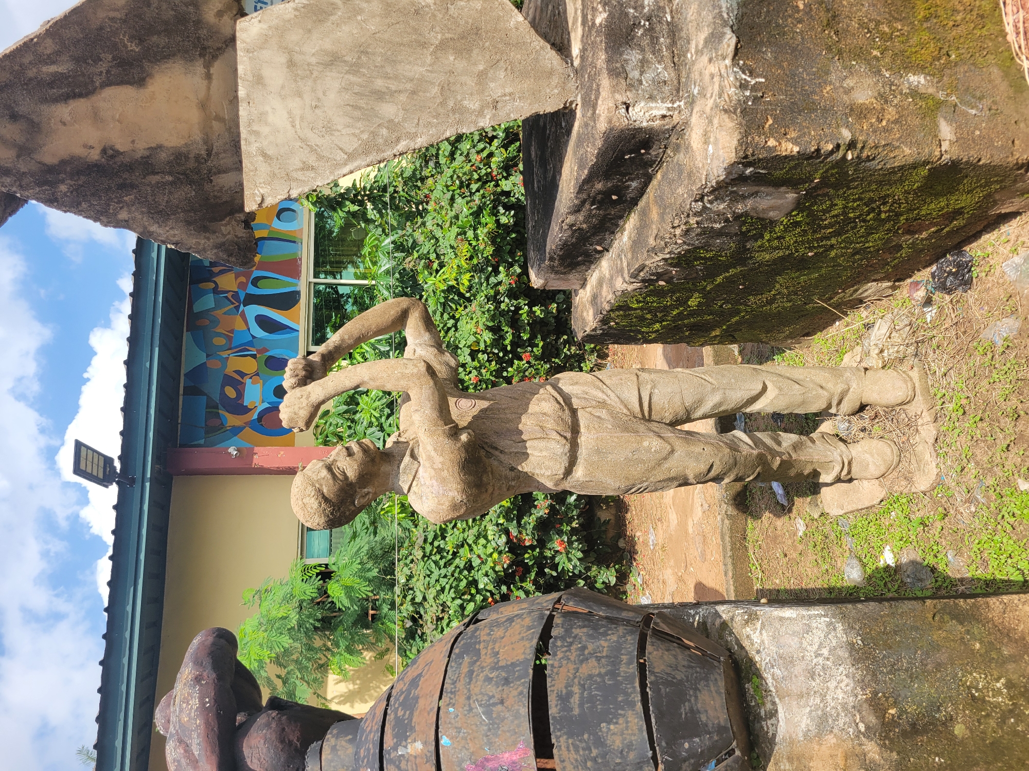 The image depicts an outdoor scene at the Egbagu, Nigeria, location. In the foreground, a light-brown, weathered statue of a man is positioned centrally. The statue, made of what appears to be concrete or stone, is depicted wearing a collared shirt and pants, with its arms raised as if holding something to its face, possibly a tool or binoculars. To the left of the statue, a large, dark, barrel-shaped sculpture with segmented metal bands is visible. To the right, a rough, concrete structure with moss growth on its surface supports a large, triangular concrete slab tilted at an angle. Behind the statue and sculptures, a dense green bush with small red flowers forms the backdrop. A building with a yellow facade, a green roof, and a colorful abstract mural is situated behind the foliage. A solar-powered light fixture is mounted on the building's overhang. The ground is uneven, with patches of grass, dirt, and scattered debris, including plastic bags. The sky above is partly cloudy with a blue hue.