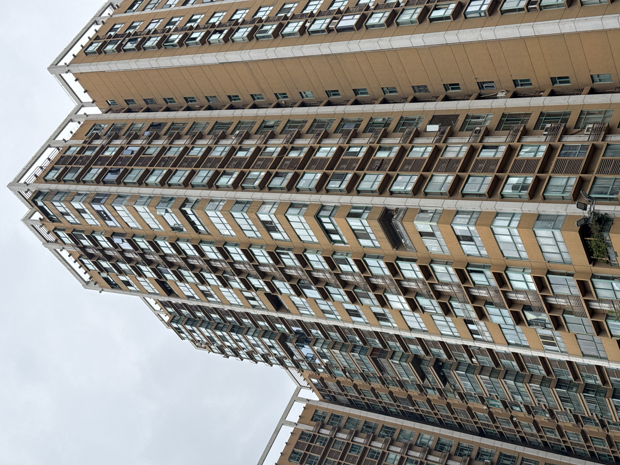 A low-angle view of a tall, brown apartment building in Jinjiang District, China. The building has many windows, most of which are open. The building appears to be residential. The sky is overcast.