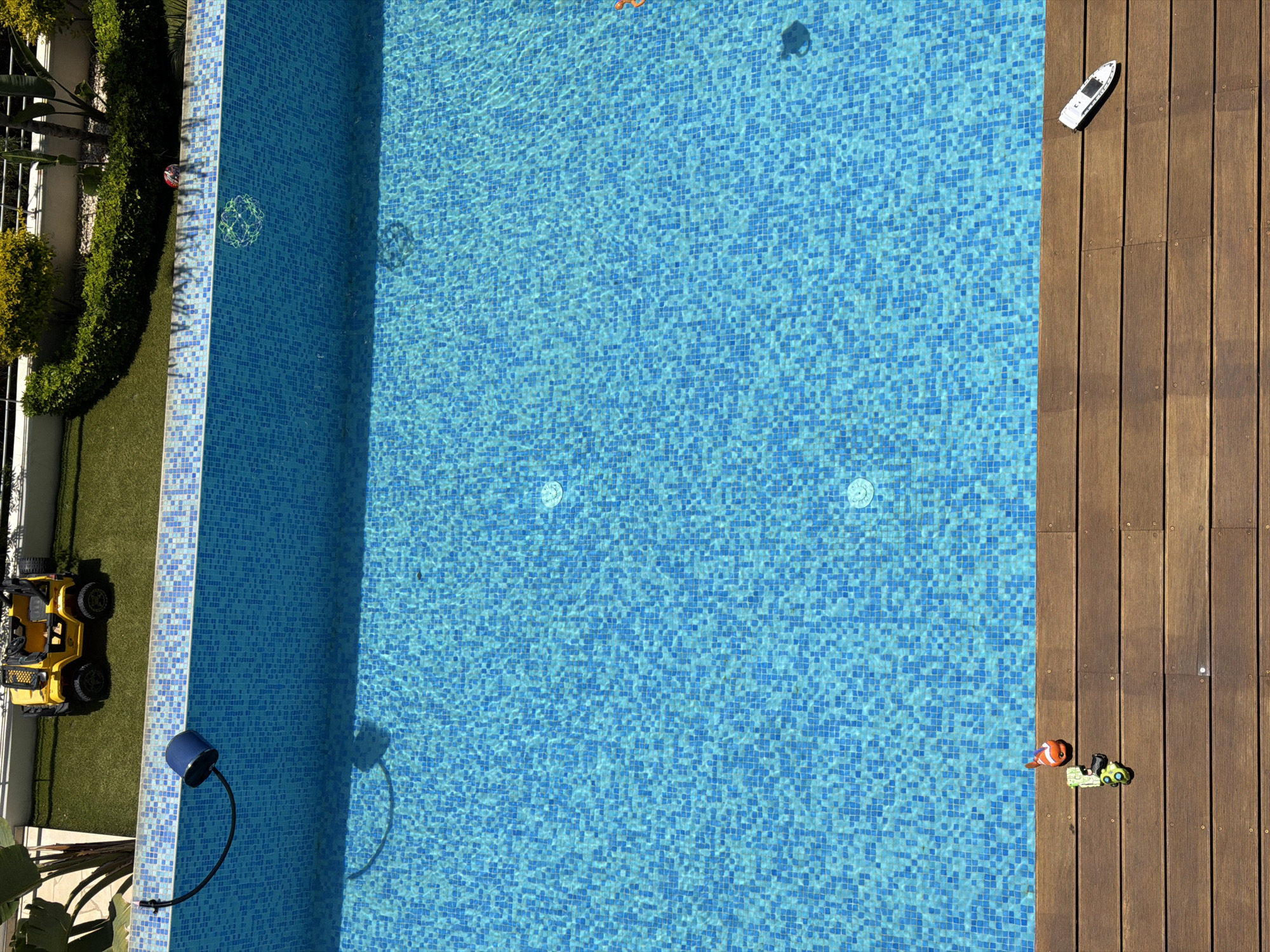 This is an aerial, eye-level shot of a swimming pool area. The pool is filled with blue tiled water and is partially bordered by a wooden deck. On the wooden deck, there are three small toys: a clownfish, a green vehicle, and a white toy boat. In the background, there is artificial turf with a yellow toy jeep parked on it, and some landscaping with bushes and plants. A grey railing can be seen above the landscaping. A blue pool cleaner is attached to the edge of the pool with a black hose. The location is the city of Agios Tychona Community, Cyprus.