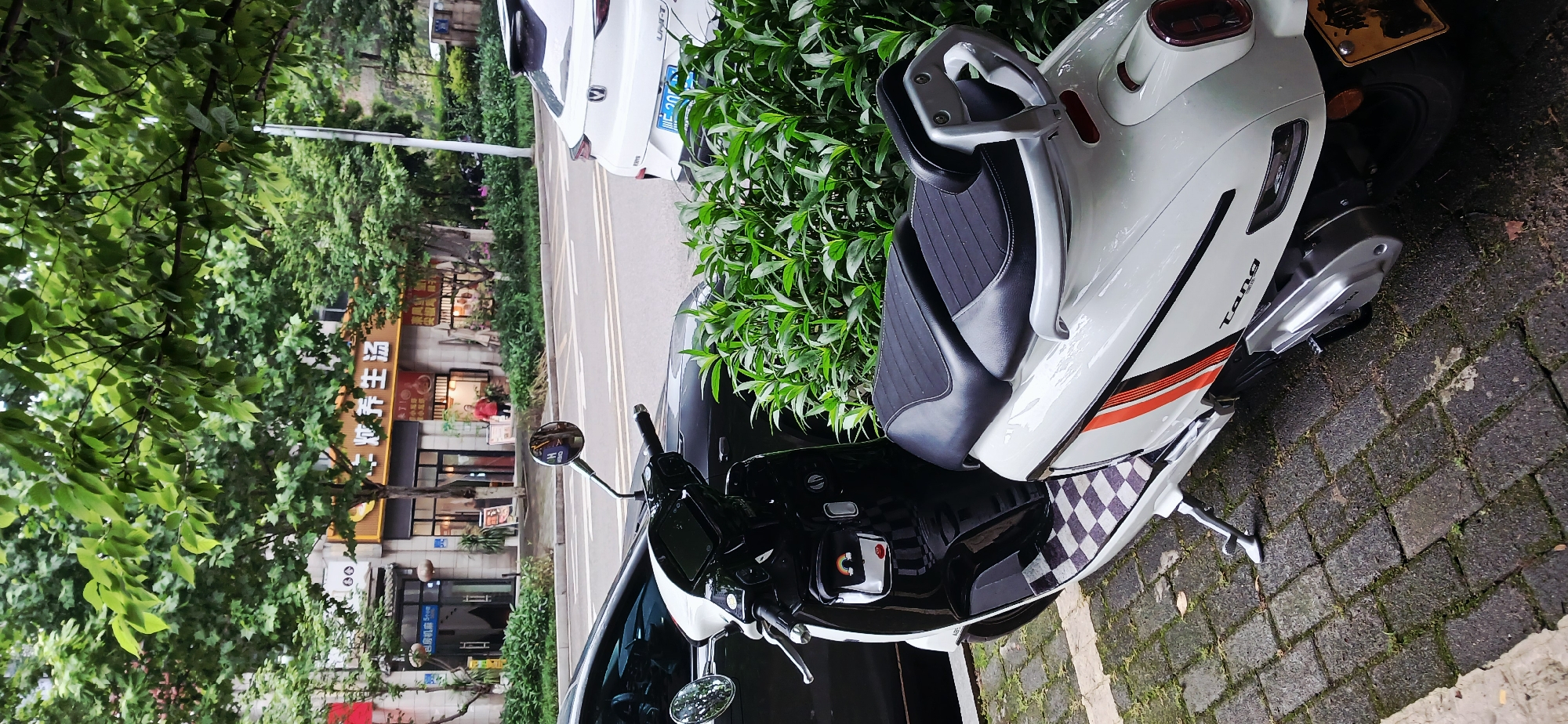 A white "Tang" scooter is parked on a gray brick walkway adjacent to a street with vegetation in Jiulongpo District, China. In the background, a restaurant with Chinese characters and a white car are visible. The scooter's seat is black, and it has an orange stripe along its side. The scooter is near greenery growing out of the ground.