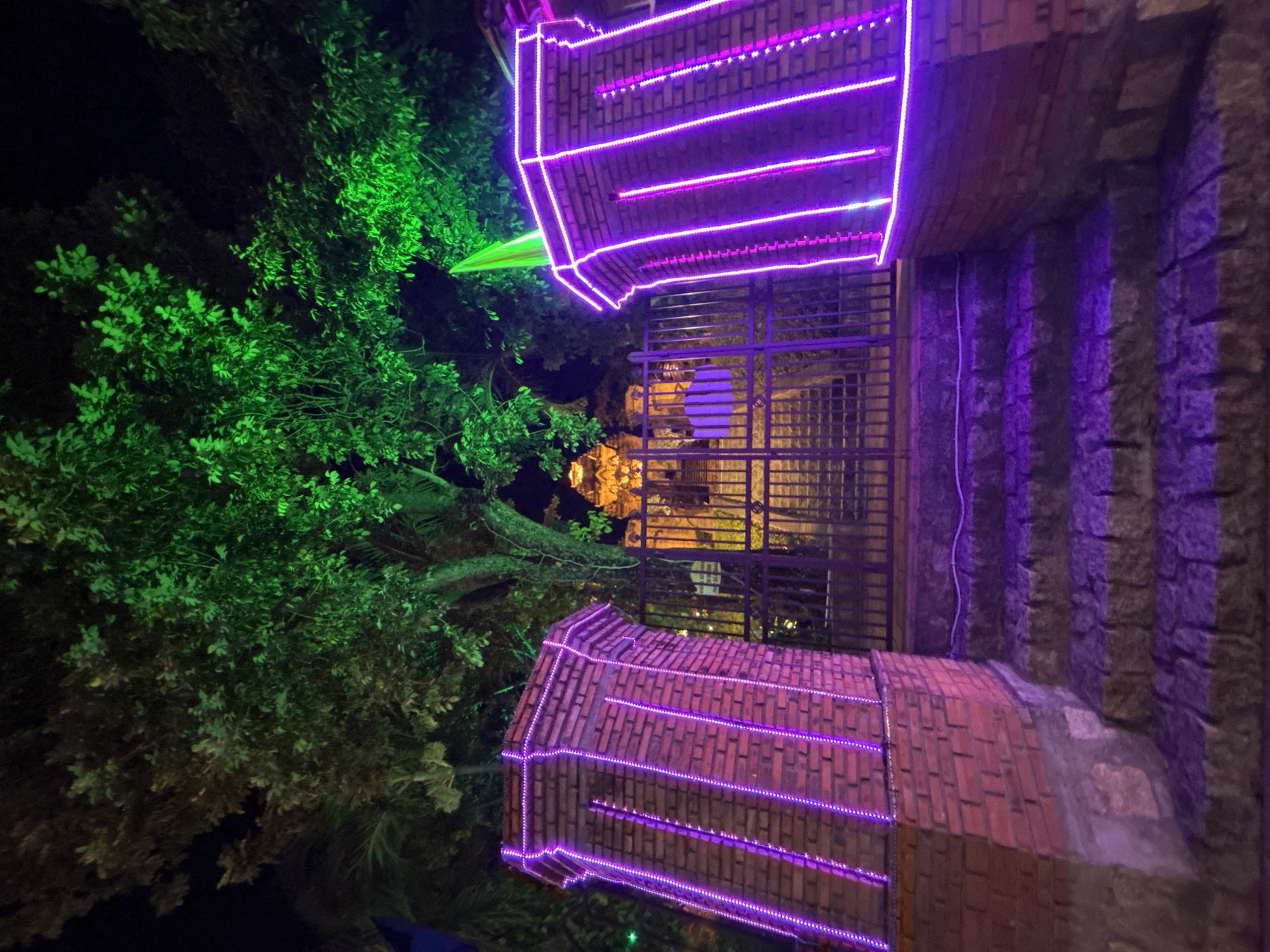 The image depicts an outdoor scene in Bắc Nha Trang, Vietnam, featuring a building with brickwork illuminated by horizontal purple LED strips. The building is viewed through a metal gate. Lush green foliage, possibly trees, is brightly lit in green hues. The scene appears to be at night, with other structures visible in the background, lit in warmer tones. Stairs leading to or from the building are visible on the right, bathed in purple light. No people are visible.