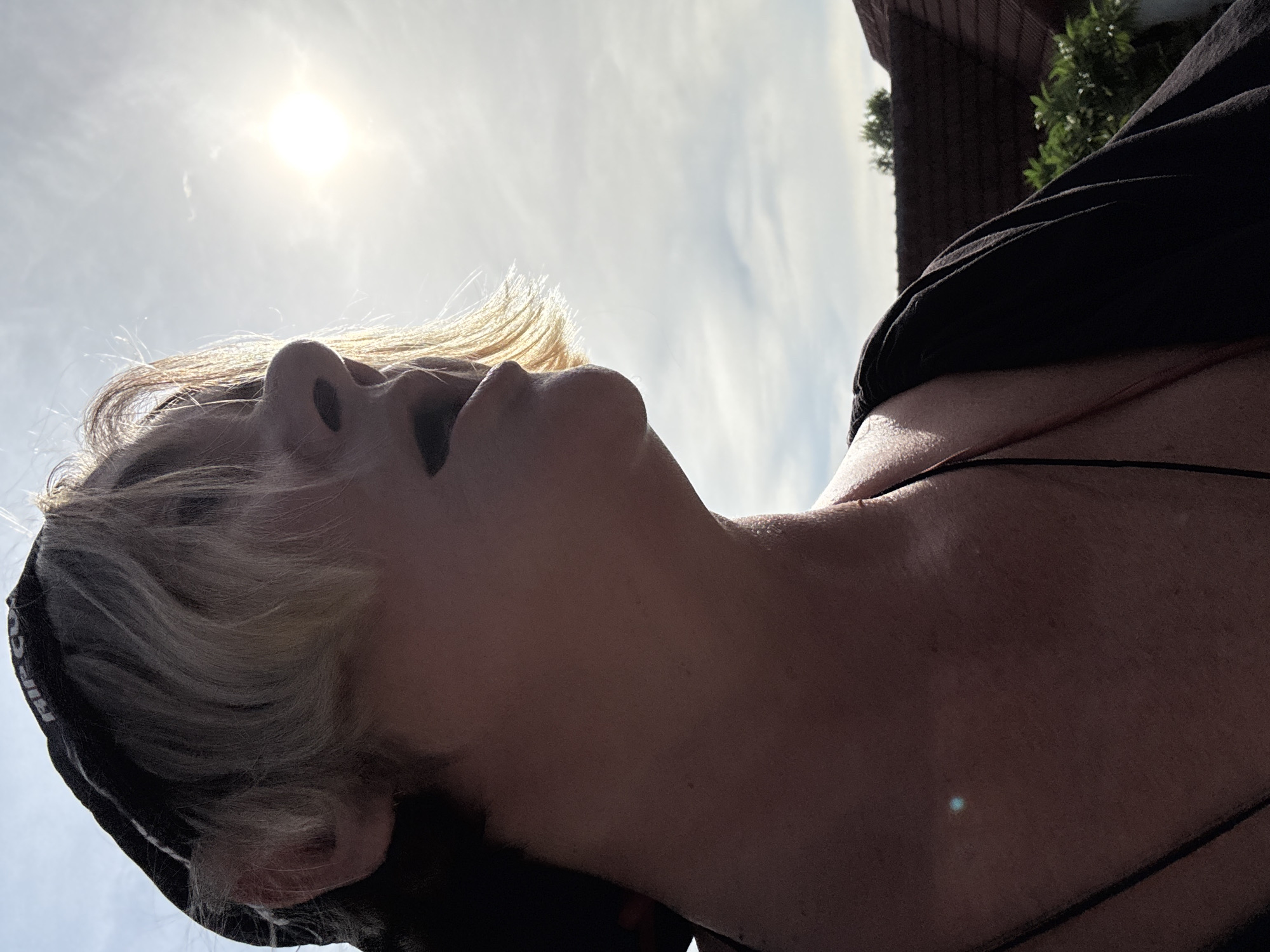 A person wearing a black baseball cap and a black shirt is looking up at the sun on a bright, cloudy day. The person's face is partially obscured by their hair, which is blonde and appears to be styled with bangs. Their mouth is slightly open. The background shows the sky, some clouds, and a few rooftops with vegetation. The image appears to have been taken outdoors in a residential area.
