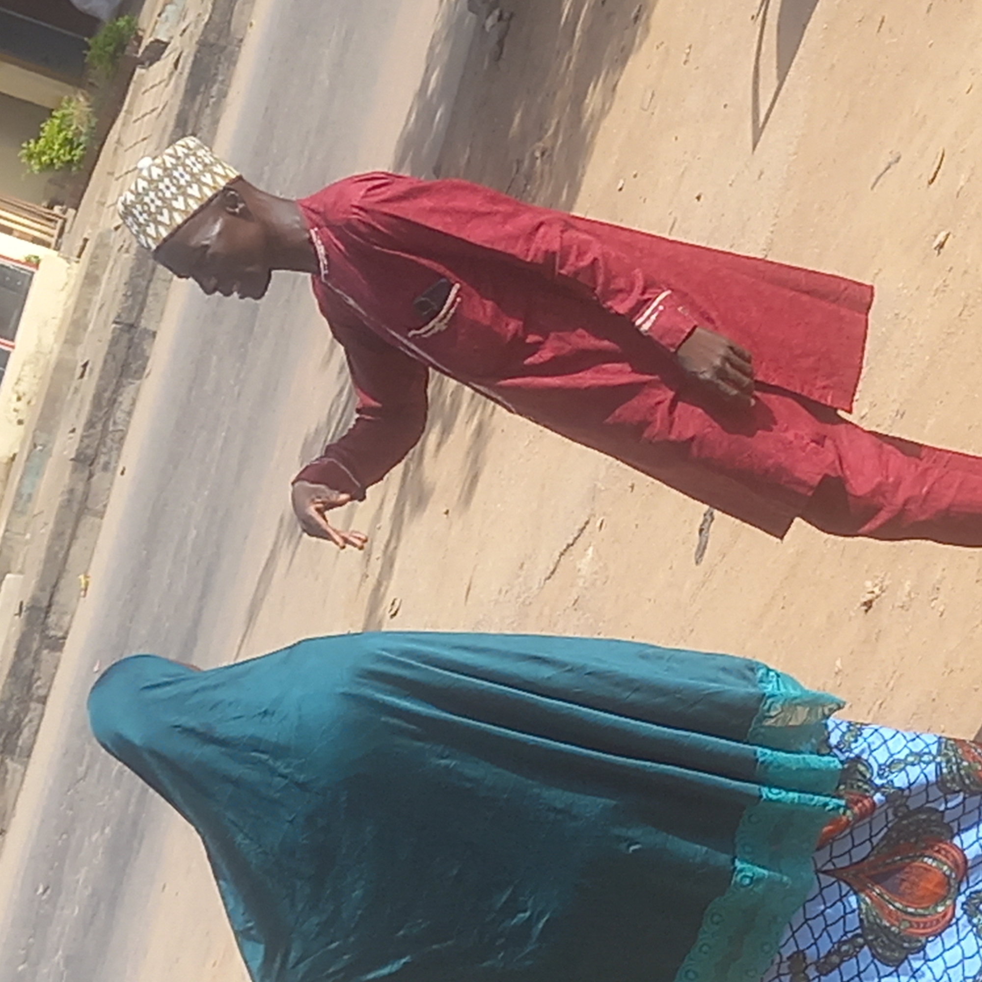 A man wearing a red garment and a traditional hat is walking on a street in Dadi Riba, Nigeria. Next to him is a woman wearing a green veil over a blue patterned fabric. The street appears to be paved and is partially shaded. In the background, there are buildings and trees.