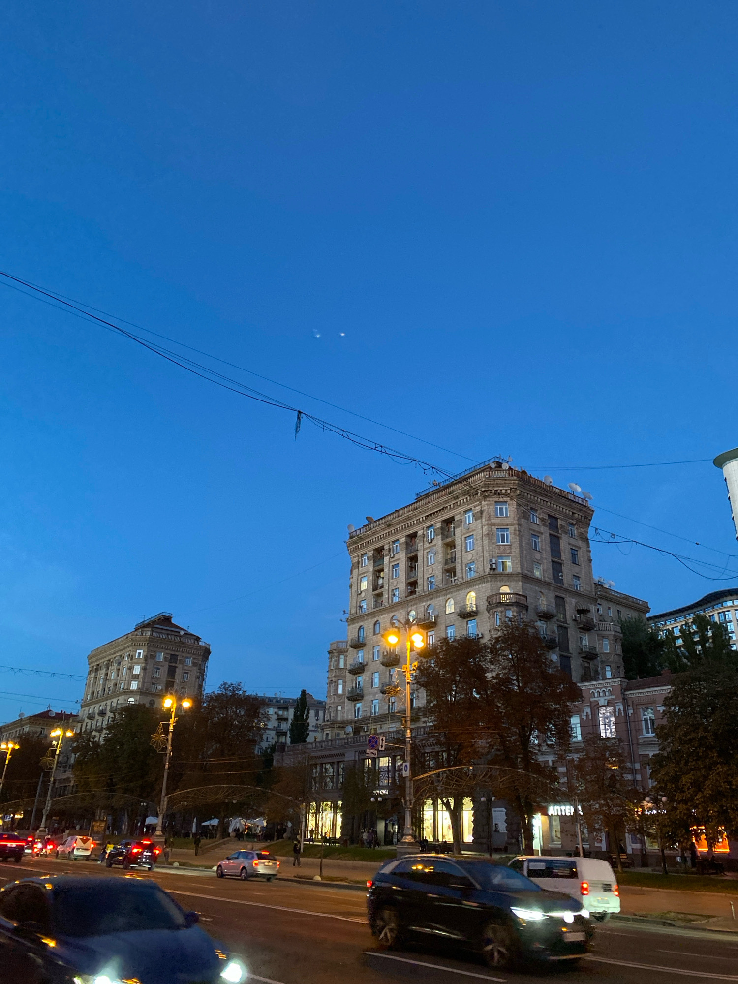 The image depicts a street scene in a city at dusk. The sky is a deep blue, with two faint celestial bodies visible near the top. Overhead, power lines crisscross the frame. In the middle ground, several large, ornate buildings with multiple stories and visible windows line the street. Some windows are illuminated, suggesting interior lights. Trees with autumn foliage are scattered throughout, and streetlights cast a warm glow. In the foreground, cars are driving on a multi-lane road. One car in the immediate foreground is dark blue and its headlights are on. Other cars, both moving and parked, are visible further down the road. A white van is also visible. The overall impression is of a bustling urban environment transitioning from day to night.