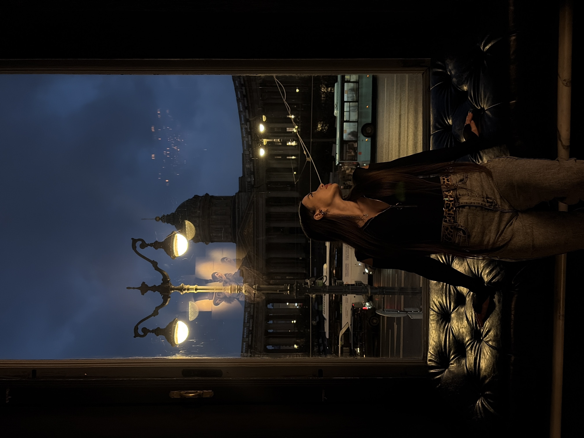 A nighttime photo captures a young woman in a dimly lit interior, leaning towards a window overlooking a street scene in Saint Petersburg, Russia. The exterior view showcases a building with architectural columns, illuminated streetlights, a city bus, and other vehicles. The reflection in the window superimposes the street scene over the woman, who is wearing a dark top and jeans with a patterned belt. She is positioned in front of a tufted leather couch. The lighting is low, emphasizing the contrast between the interior shadows and the brighter, illuminated scene outside the window.