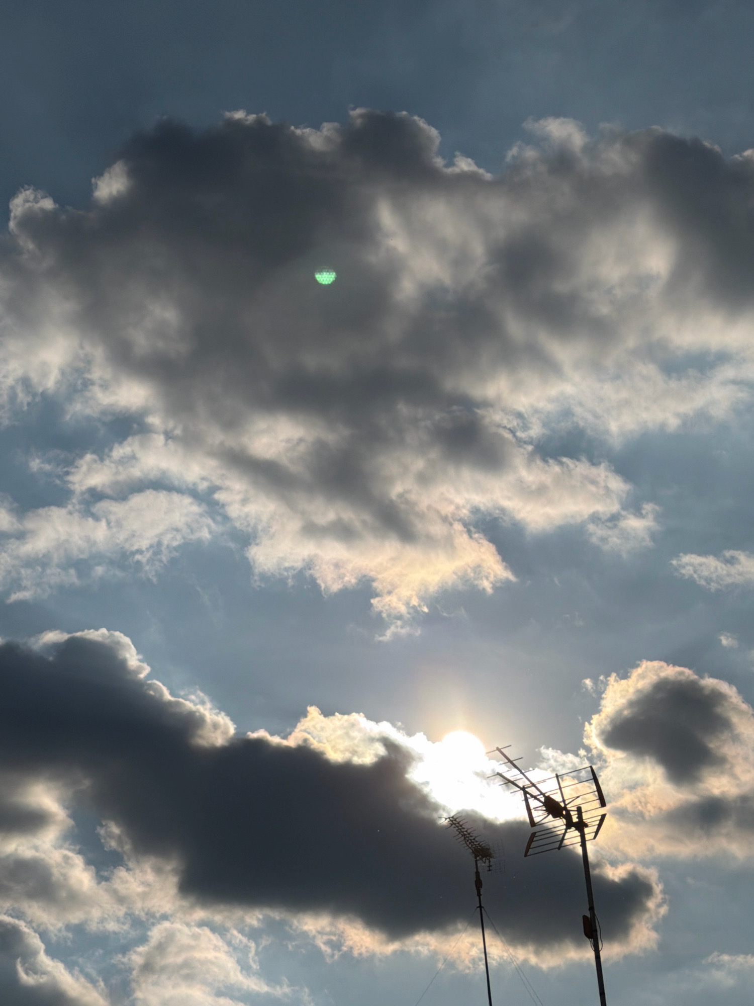 A digital photograph shows a cloudy sky with the sun peeking through. Two television antennas are silhouetted against the bright sky. The antenna on the right is larger and more complex than the one on the left. The clouds are varied in texture and density, with some appearing dark and heavy while others are lighter and more wispy. A small, green, circular object with a grid pattern is visible within one of the darker clouds in the upper left quadrant of the image. The sun is a bright, hazy orb, partially obscured by clouds and antennas.