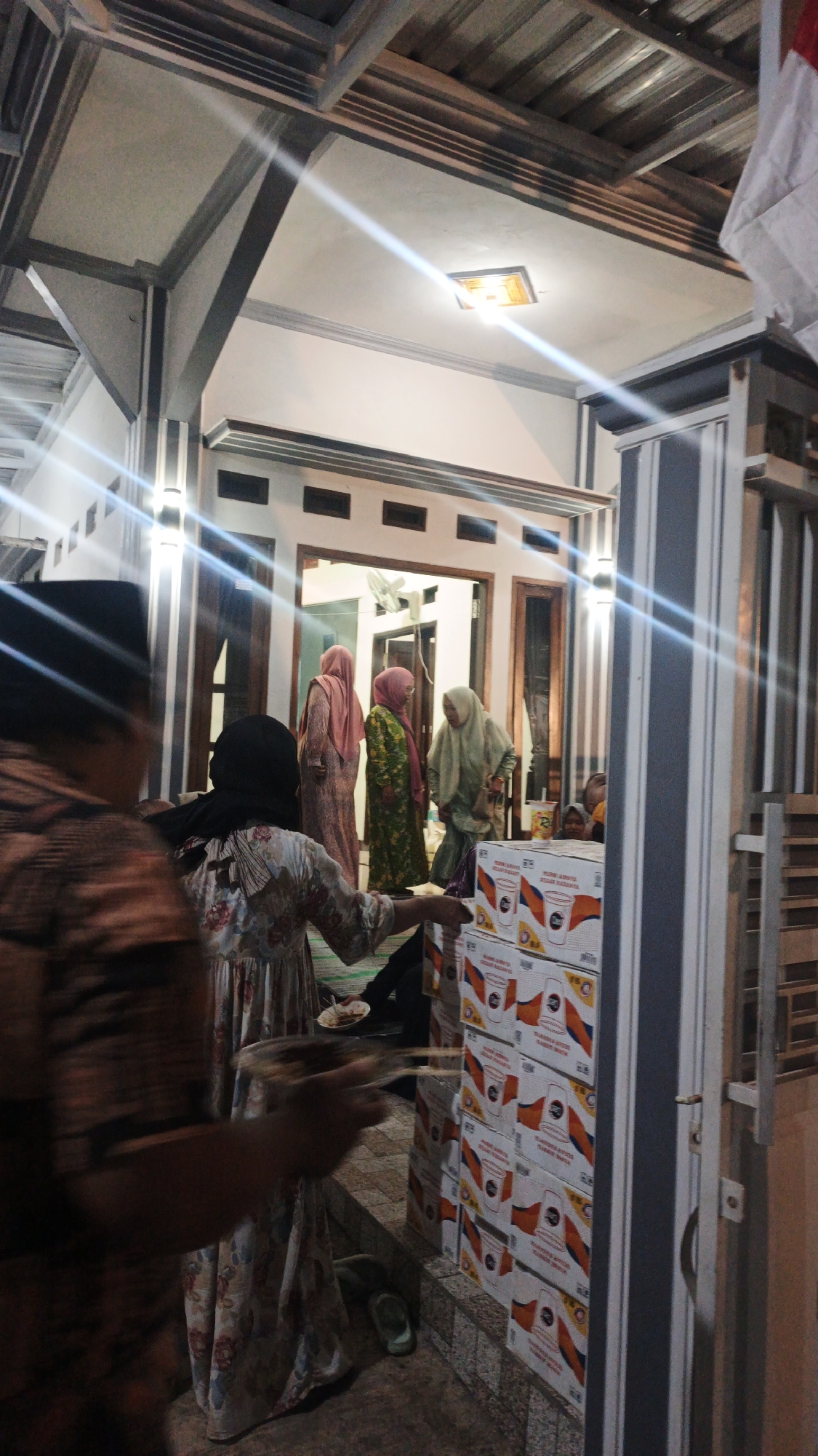 A group of people are gathered outside a building at night. The building appears to be a house with a covered porch area. A person in the foreground, facing away from the camera, is holding a plate with food. To their right, a stack of boxes with an orange and white logo is visible. Inside the building, through an open doorway, several women wearing headscarves are standing. One woman in the middle is wearing a green patterned dress, and another to her right is in a light green headscarf. Another woman to the far left wears a pink headscarf and a long pink dress. The environment is dimly lit by overhead lights and some ambient light emanating from the building. The image appears to have been taken in Pandanarum, Indonesia, given the context provided.