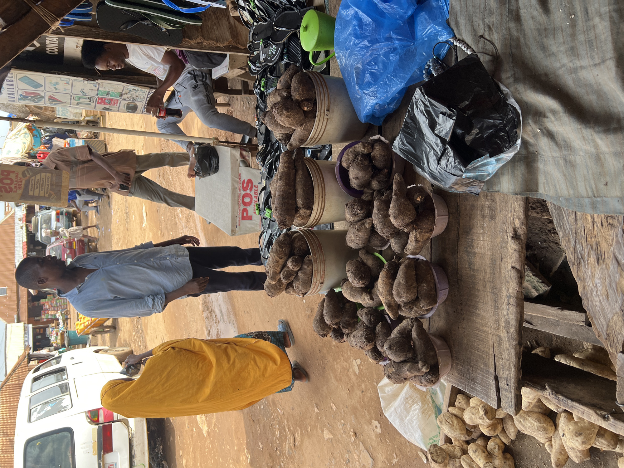 The image depicts an outdoor market scene in Abuja, Nigeria. Buckets and baskets are overflowing with brown root vegetables, likely yams, displayed on a wooden table. A man in a light blue shirt and dark pants stands near the table. A woman wearing a yellow garment is also present. In the background, a white van and other vehicles are visible. Other individuals can be seen walking and standing nearby. The ground is reddish-brown, suggesting a dirt or unpaved surface. Above the root vegetables are various items, including shoes, a blue plastic bag, and a black plastic bag.