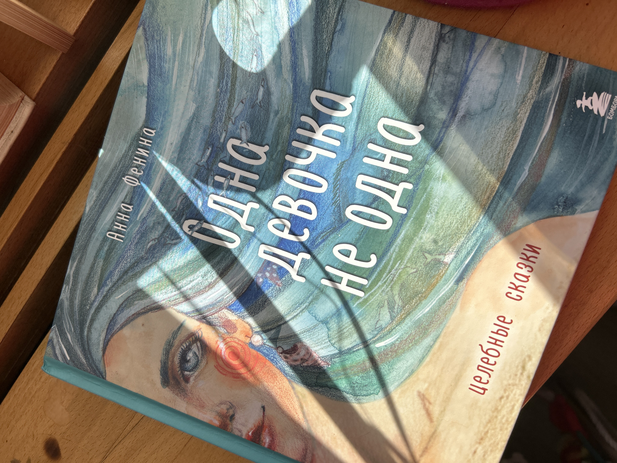 The image shows a book lying on a wooden surface. The book's cover features a painting of a woman with blue and green hues suggesting water or a dreamlike state. The title, in Russian, is prominently displayed in white, and the author's name is written vertically on the left side. The words "целебные сказки" are printed in red. A logo for the publisher is in the top right corner. Sunlight creates shadows on the book's cover. The environment is implied to be indoors, and the wooden surface looks like furniture. The city of Moscow, Russia is not mentioned in the image.