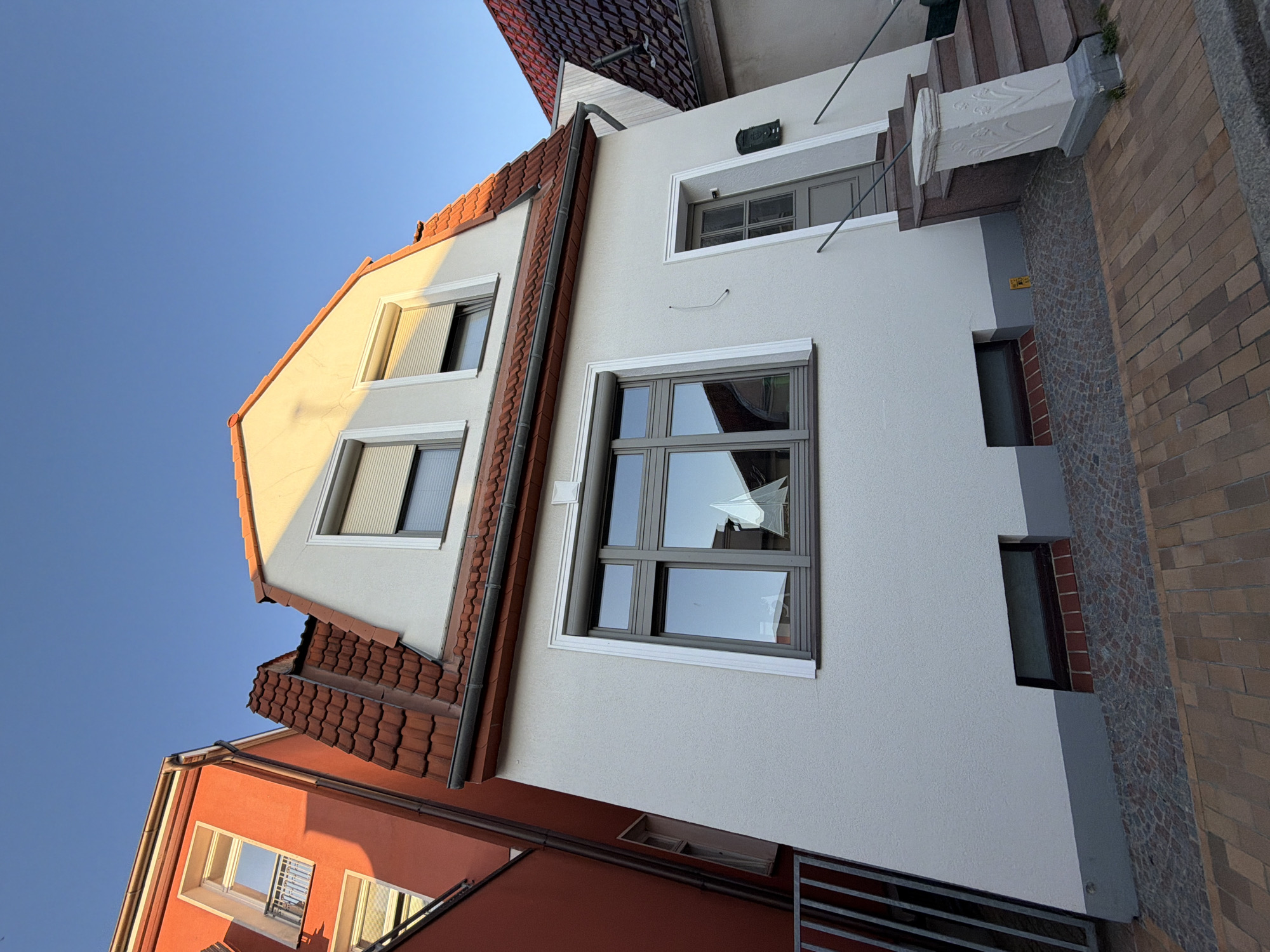 The image displays a street view in Rostock, Germany, featuring the facade of a multi-story residential building. The building has a light-colored stucco exterior with a red-tiled, steeply pitched roof. There are several windows visible, including a large, multi-pane window on the ground floor reflecting a sailboat model. Above this, on the upper level, are two smaller windows with roll-down shutters. A gray-painted door is located to the right of the ground-floor window, accessible by a set of granite steps. A green mailbox is mounted to the right of the door. The foreground shows a paved sidewalk made of reddish-brown bricks and gray cobblestones. Adjacent buildings, one painted bright orange, are also partially visible, creating a dense urban setting. The sky is clear and blue, with the sun casting warm light on the building.