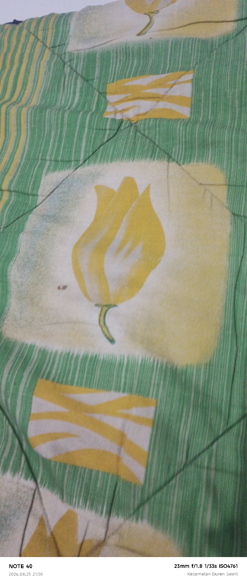 The image is a close-up, overhead view of a patterned fabric, likely a blanket or comforter. The fabric features a repeating pattern of green and white vertical stripes interspersed with yellow and white rectangular motifs and yellow tulip-like floral designs. The pattern is quilted, with visible stitching lines creating diamond shapes across the fabric. The lighting appears to be artificial, casting some shadows and creating a slightly uneven illumination. The bottom of the image includes text overlay which reads "NOTE 40", "2026.08.25 21:30", "23mm f/1.8 1/33s ISO4761", and "Kecamatan Duren Sawit".
The text "Kecamatan Duren Sawit" indicates the location is the Duren Sawit subdistrict, which is part of the city of East Jakarta, Indonesia.