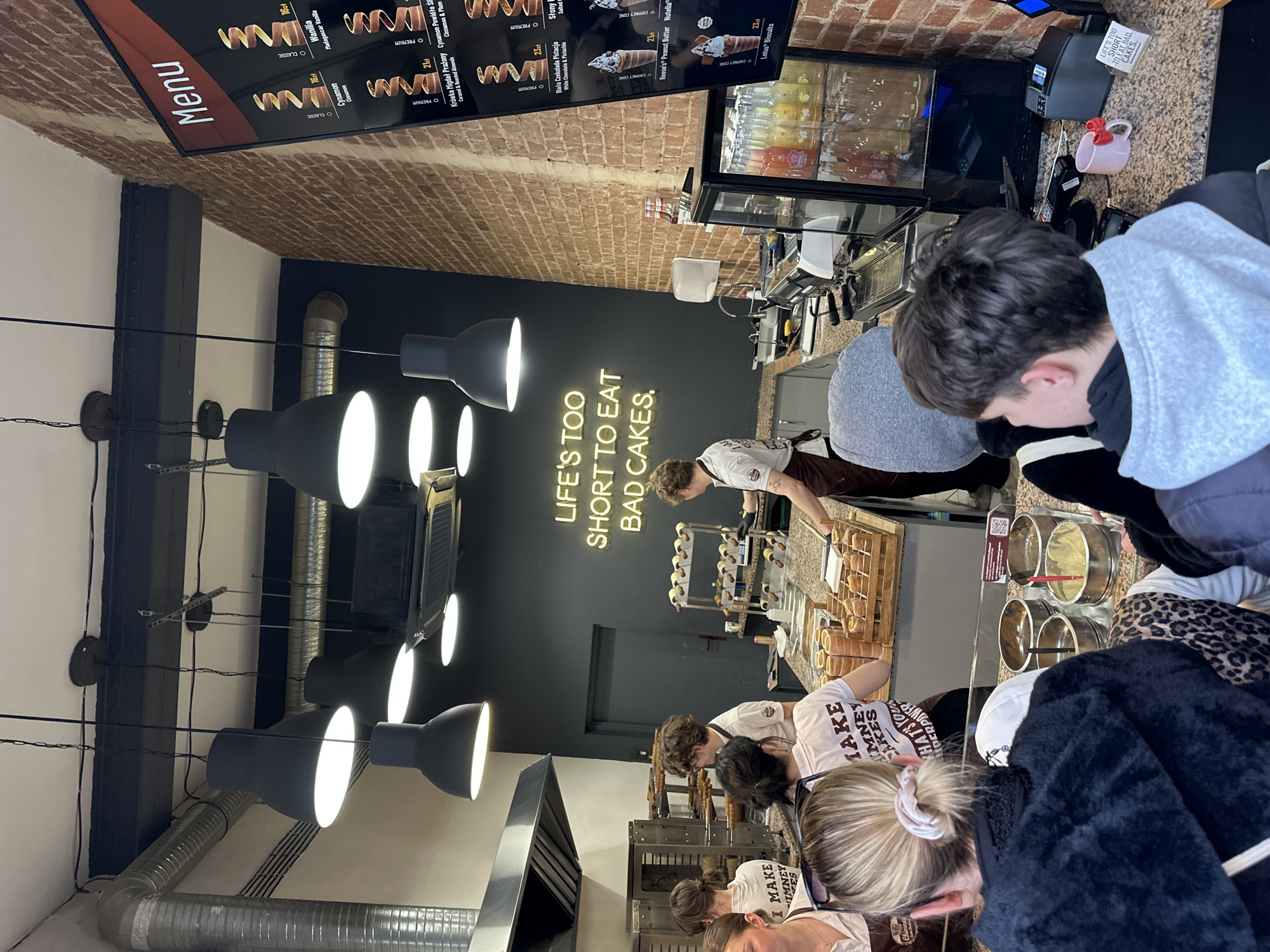 The image captures the interior of Kurtos Kolacs, a chimney cake bakery in Wrocław, Poland. A queue of patrons is seen waiting to order, with at least eight individuals visible. A menu board displaying cake options and prices is positioned on the brick wall above the counter. Employees in white shirts and brown aprons work behind the counter, preparing and serving the cakes. The bakery features industrial-style lighting fixtures and a neon sign stating, "Life's Too Short To Eat Bad Cakes." The counters display the available toppings and prepared chimney cakes. A refrigerated display case contains beverages.