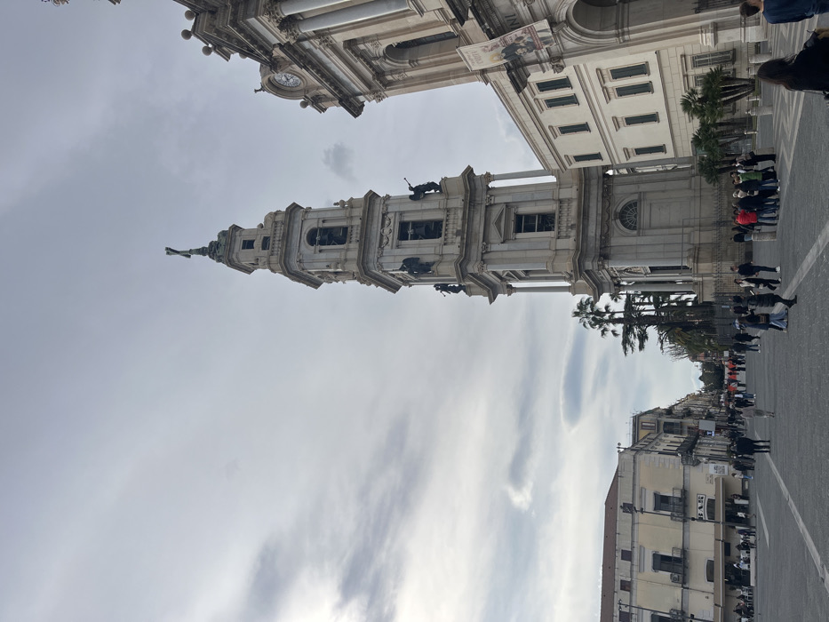 This image depicts a wide-angle view of the Piazza Bartolo Longo in Pompei, Italy, focusing on the Bell Tower and a portion of the Pontifical Shrine of the Blessed Virgin of the Rosary of Pompei.
In the center-right, the prominent, multi-tiered bell tower of the basilica rises, constructed from light-colored stone. It features several levels with arched openings, columns, and statuary. On an upper tier, two dark-colored statues of figures appear to be holding trumpets, while the tower culminates in a cross. To the immediate right of the bell tower, a section of the basilica's main facade is visible, displaying classical architectural elements, a circular clock near its upper portion, and a vertical banner with text and images. Palm trees are visible at the base of the bell tower and alongside the basilica.
The foreground and left side of the image show a large, paved public square (piazza) populated by numerous pedestrians. Individuals and small groups are dispersed across the square, walking, standing, and congregating. Some people are gathered closer to the basilica entrance. On the far left, multi-story buildings with commercial storefronts, including one labeled "BAR," line the edge of the piazza. The sky is overcast with varying cloud formations.