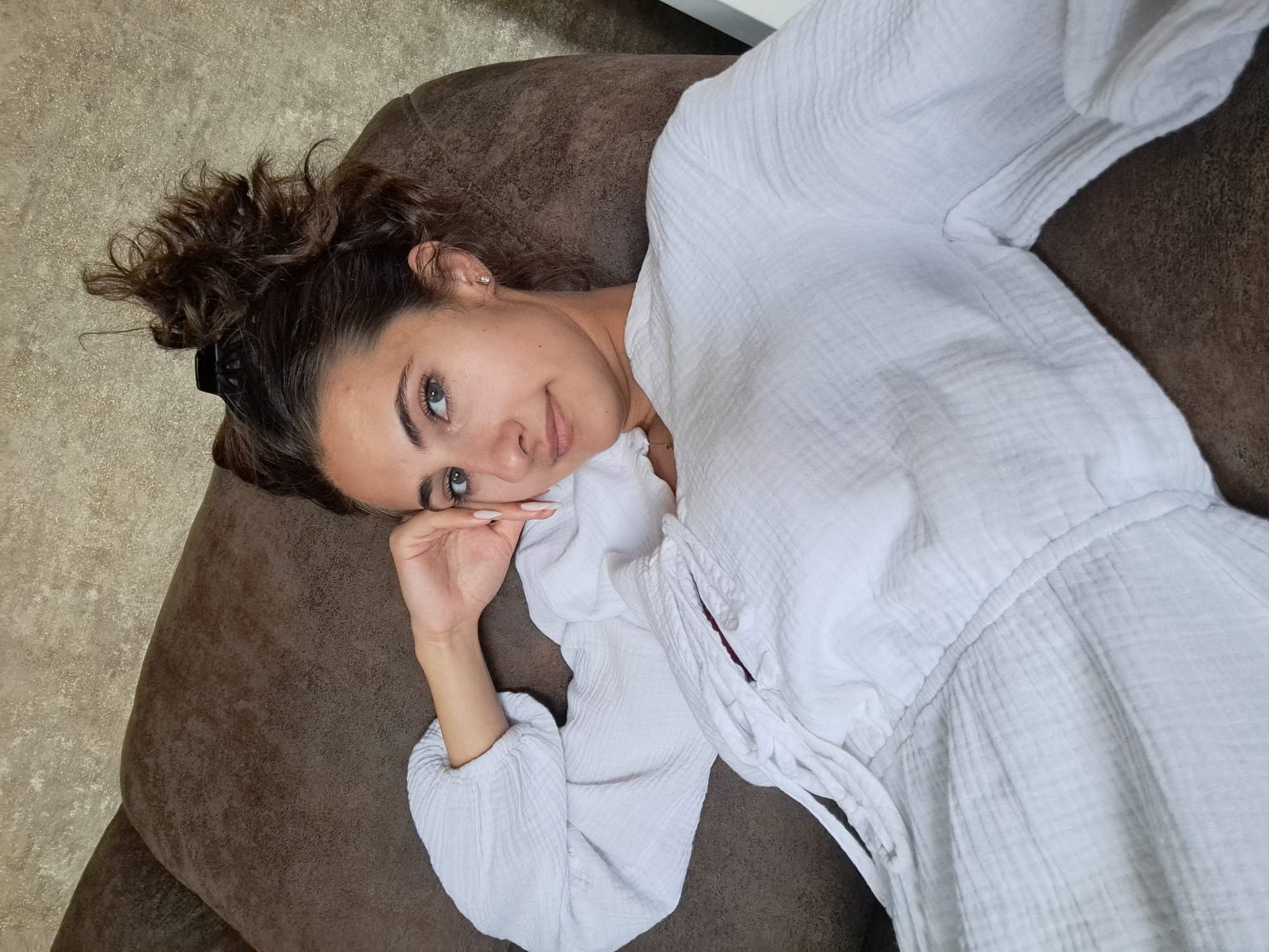 This media file is a selfie taken from a high-angle perspective of a young woman lying on a brown couch. She has dark, curly hair pulled back and is wearing a white gauze-like tunic. Her eyes are open and looking towards the camera, with a gentle smile on her lips. Her left hand is raised to her cheek, and her fingernails are painted a light color. The background consists of the brown couch and a textured, light-colored floor. The lighting suggests it is daytime, possibly indoors given the couch. The overall mood is relaxed and casual. There is no visible text in the image.