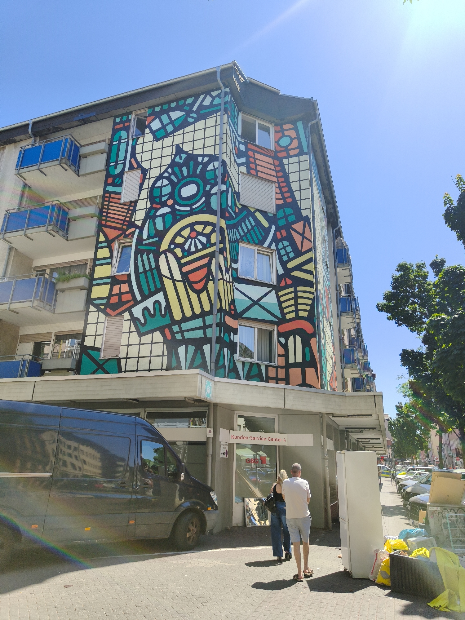 The image depicts a street scene in Mannheim, Germany, featuring a building adorned with a large, colorful mural. The mural has an abstract design with geometric shapes and patterns in various bright colors, including yellow, orange, teal, and black, covering a significant portion of the building's facade. Below the mural, the building appears to be a commercial or residential complex with balconies.
In the foreground, a dark-colored van is parked along the sidewalk. A rainbow-colored light flare is visible across the van and the sidewalk, indicating bright sunlight. Two individuals, a man and a woman, are walking on the sidewalk. The man is wearing a white t-shirt and light-colored shorts, while the woman is wearing a dark top and jeans.
Adjacent to the building's entrance, there are several large items placed on the sidewalk, including a white refrigerator, yellow trash bags, and other discarded items, suggesting possible moving or clean-up activity. A sign above the entrance reads "Kunden-Service-Center 4".
The street continues to the right, lined with parked cars and trees. The sky is clear and blue, with visible sunlight creating lens flare. The overall scene suggests a sunny day in an urban environment.