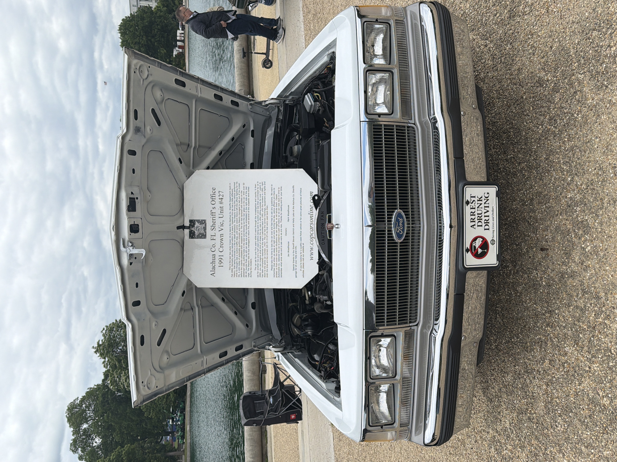 A white 1991 Crown Victoria, likely a police car given its markings, sits parked on a stone paved area in Washington, D.C. The hood is open, revealing the engine and a sign with the car's history and other information. An "ARREST DRUNK DRIVING" license plate is visible on the front bumper. In the background, a body of water, possibly the Potomac River, is partially visible with some trees and building. An adult male is standing near the car, dressed in dark jacket, pants, and sneakers. The sky overhead is mostly overcast, and a corner of a building is visible on the left. A black foldable camping chair is located next to the car, on the left.